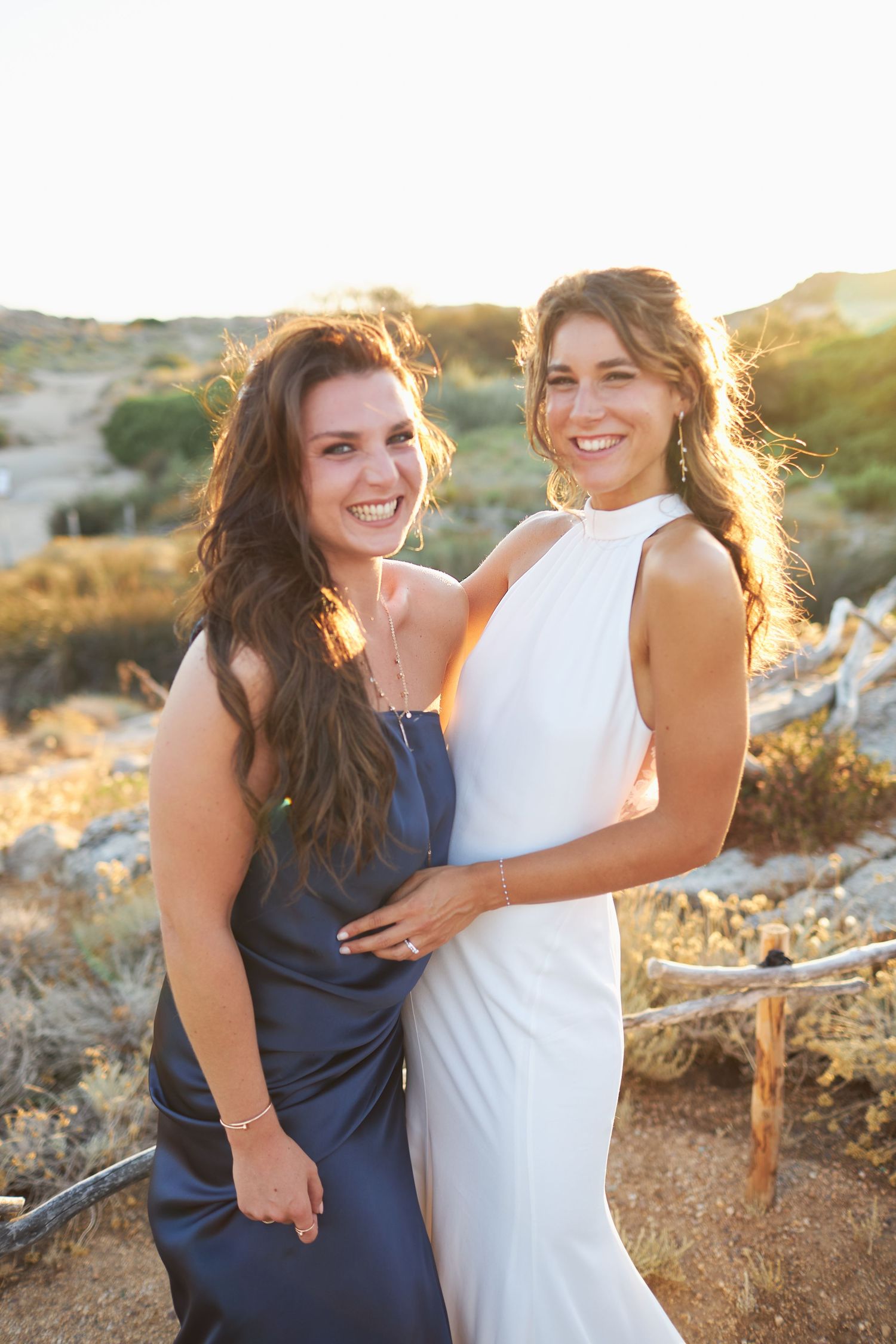 Bride in white halter gown smiling with bridesmaid in navy dress at golden hour outdoor wedding.