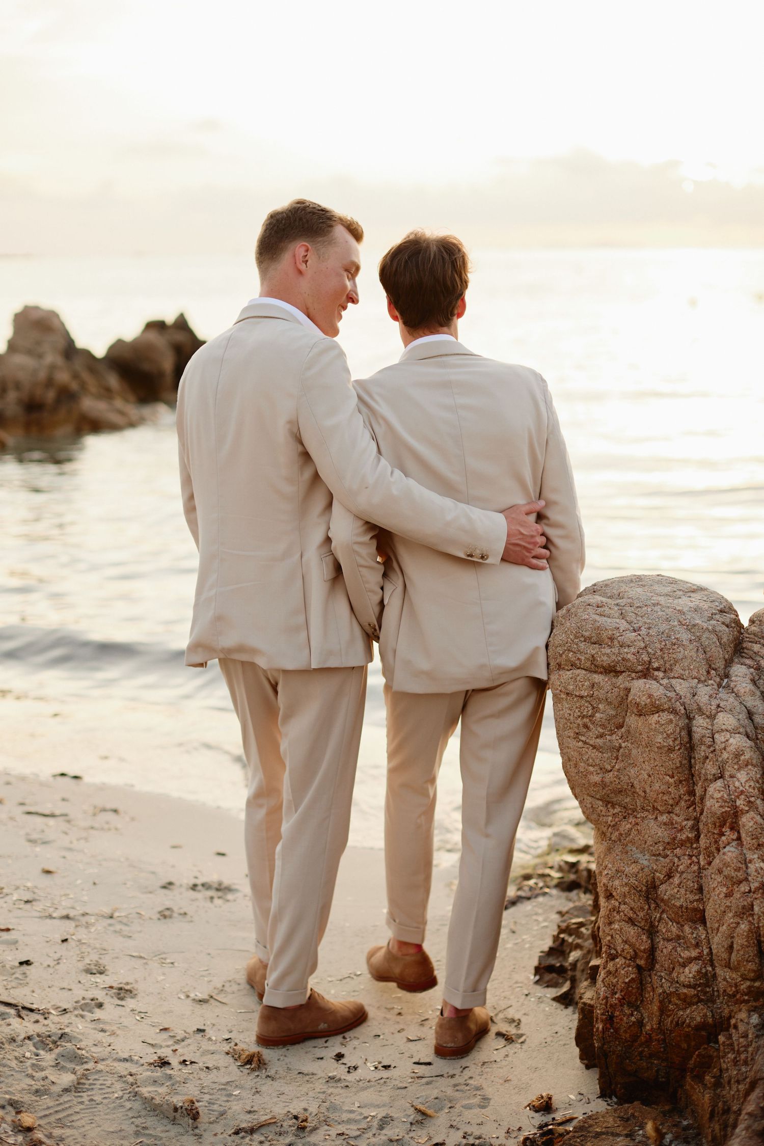 Two grooms in matching beige suits embracing on a sandy beach at sunset by the ocean.