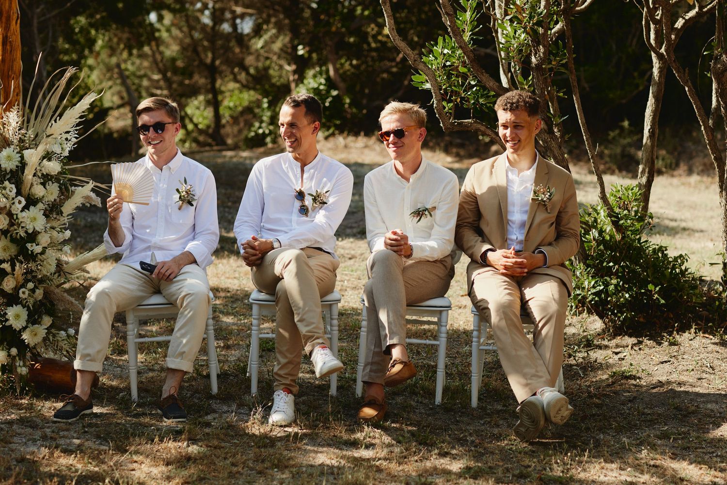 Four young men in white shirts and beige pants hold programs at an outdoor wedding ceremony.
