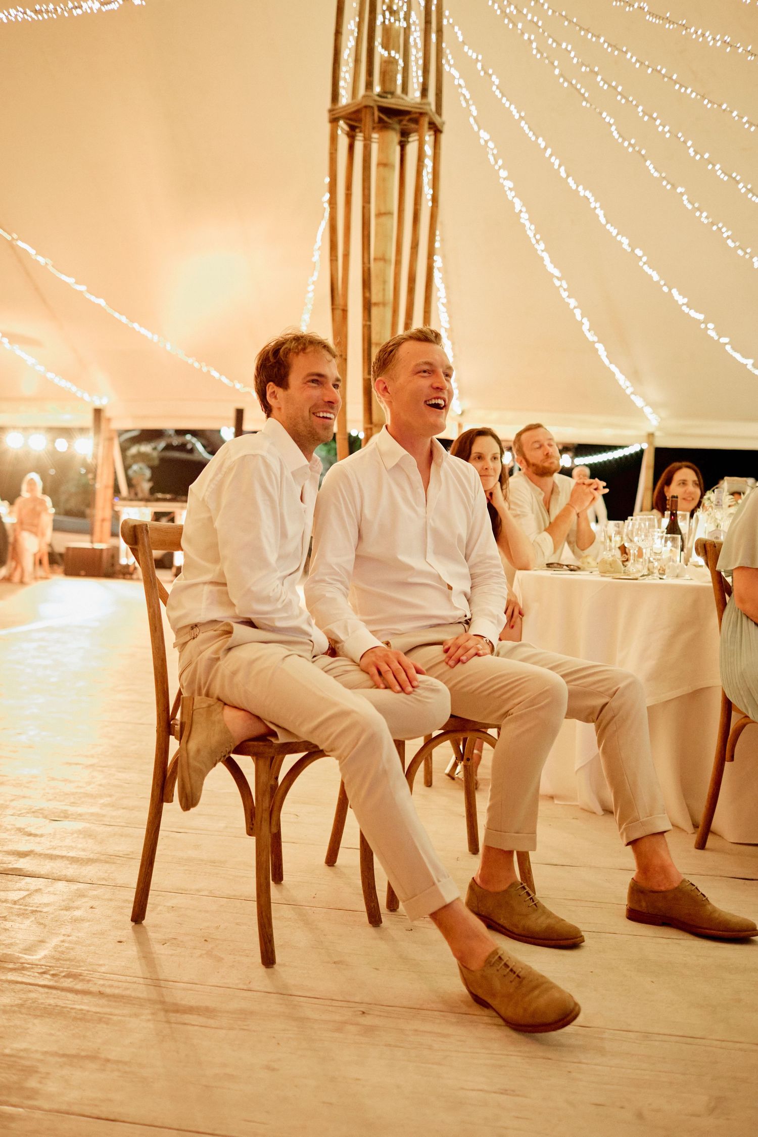 Two grooms laughing joyfully while seated at their wedding reception under fairy lights in an elegant tent.