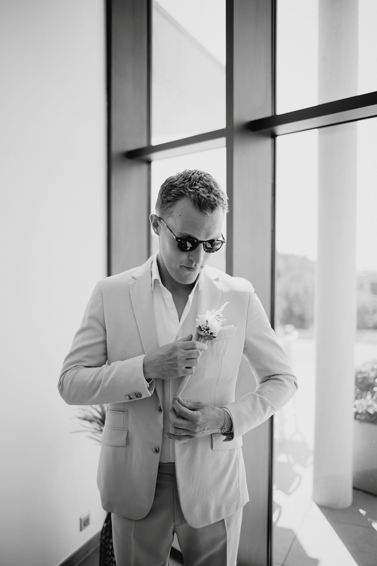 Groom in beige suit and sunglasses adjusting boutonniere in black and white portrait by window.