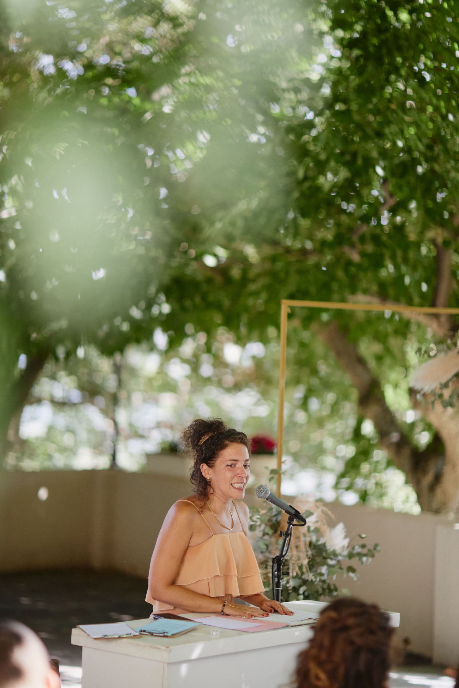 Smiling woman in peach dress speaking at outdoor wedding ceremony podium surrounded by lush green trees.