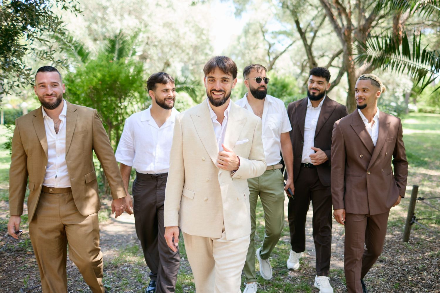 Groom in cream suit walks confidently with groomsmen in brown suits through a green garden.