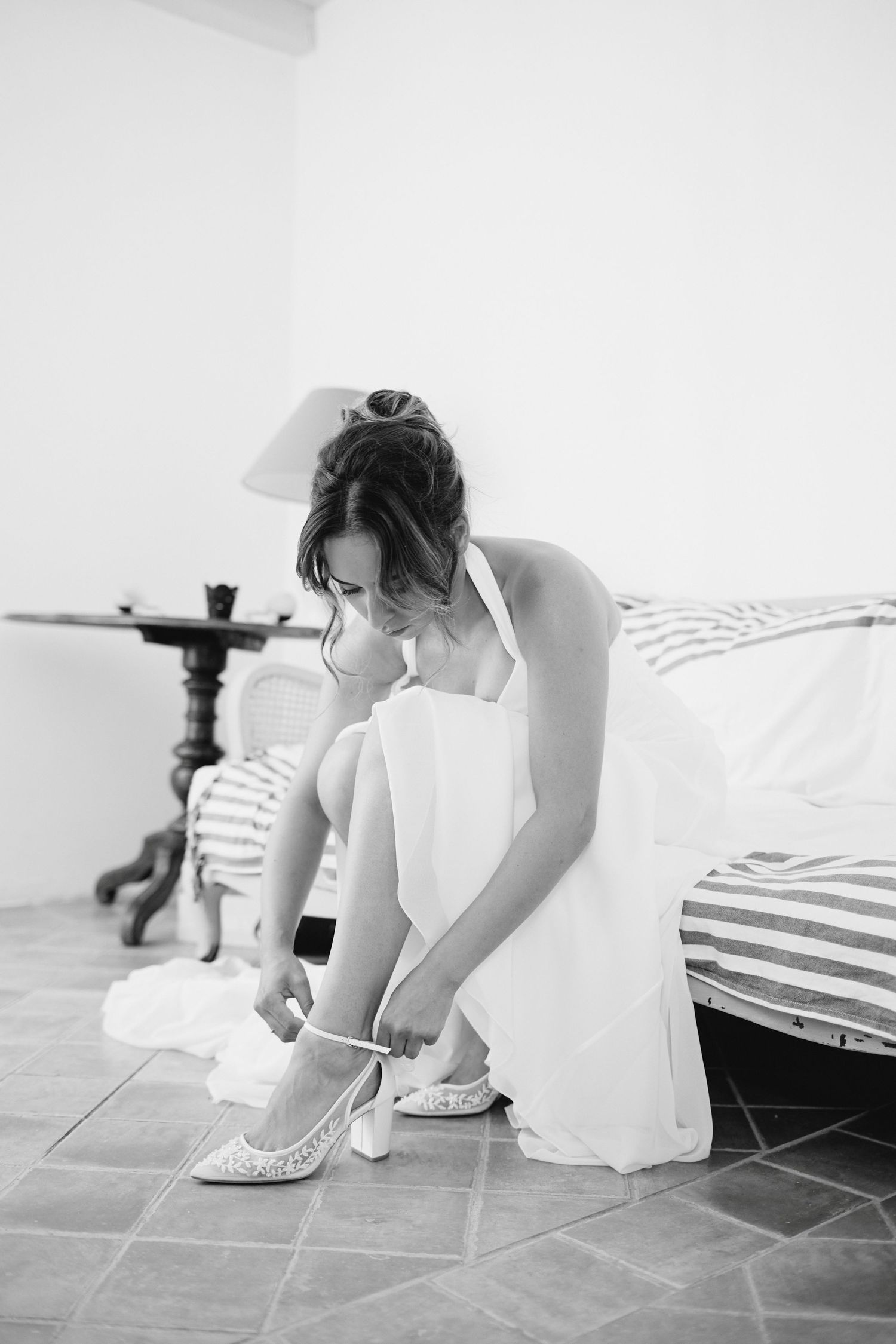 Black and white photo of bride in white halter gown putting on lace wedding shoes before ceremony.