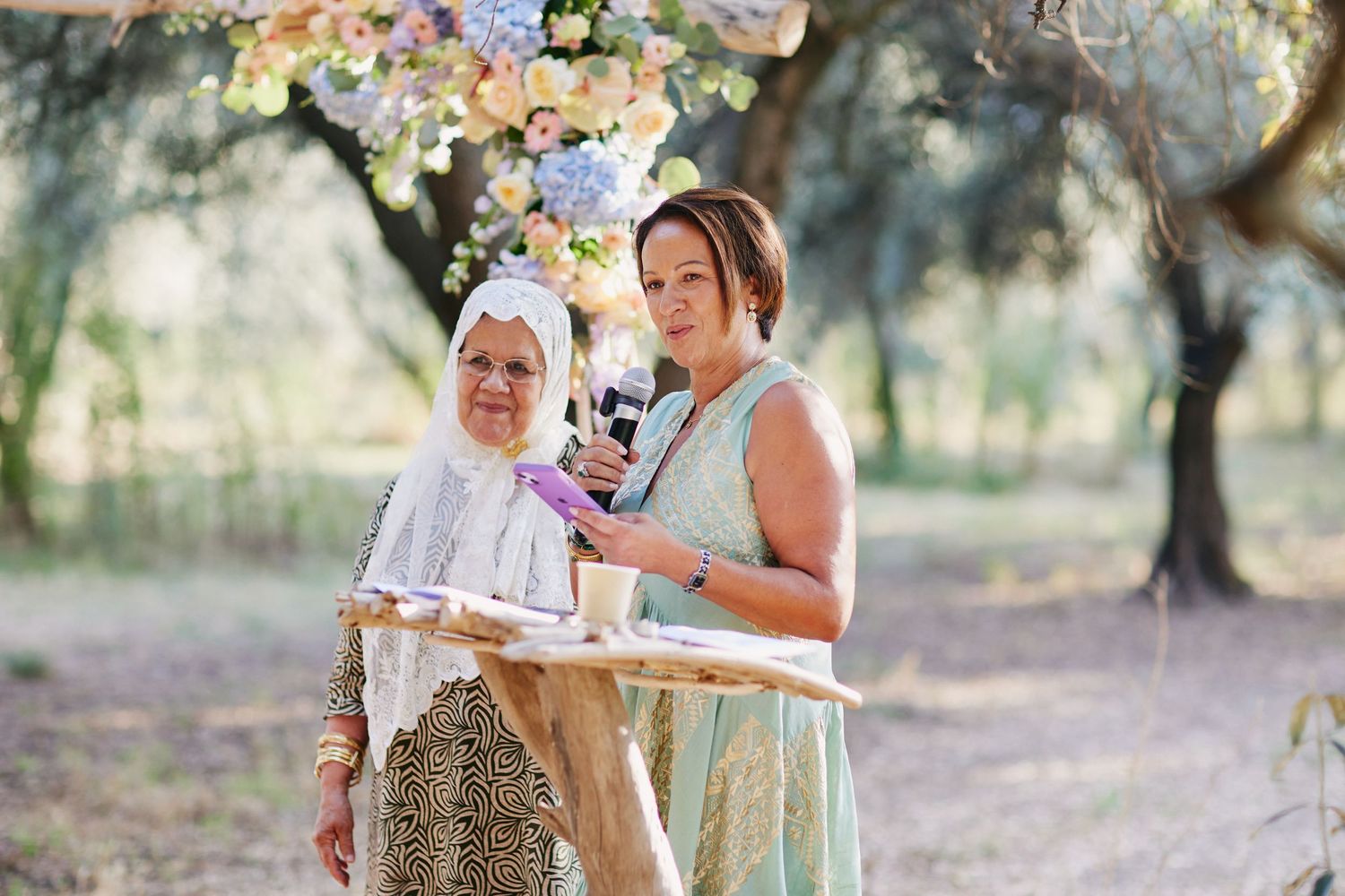 Two women stand at a floral wedding altar, one holding a tray during an outdoor ceremony.
