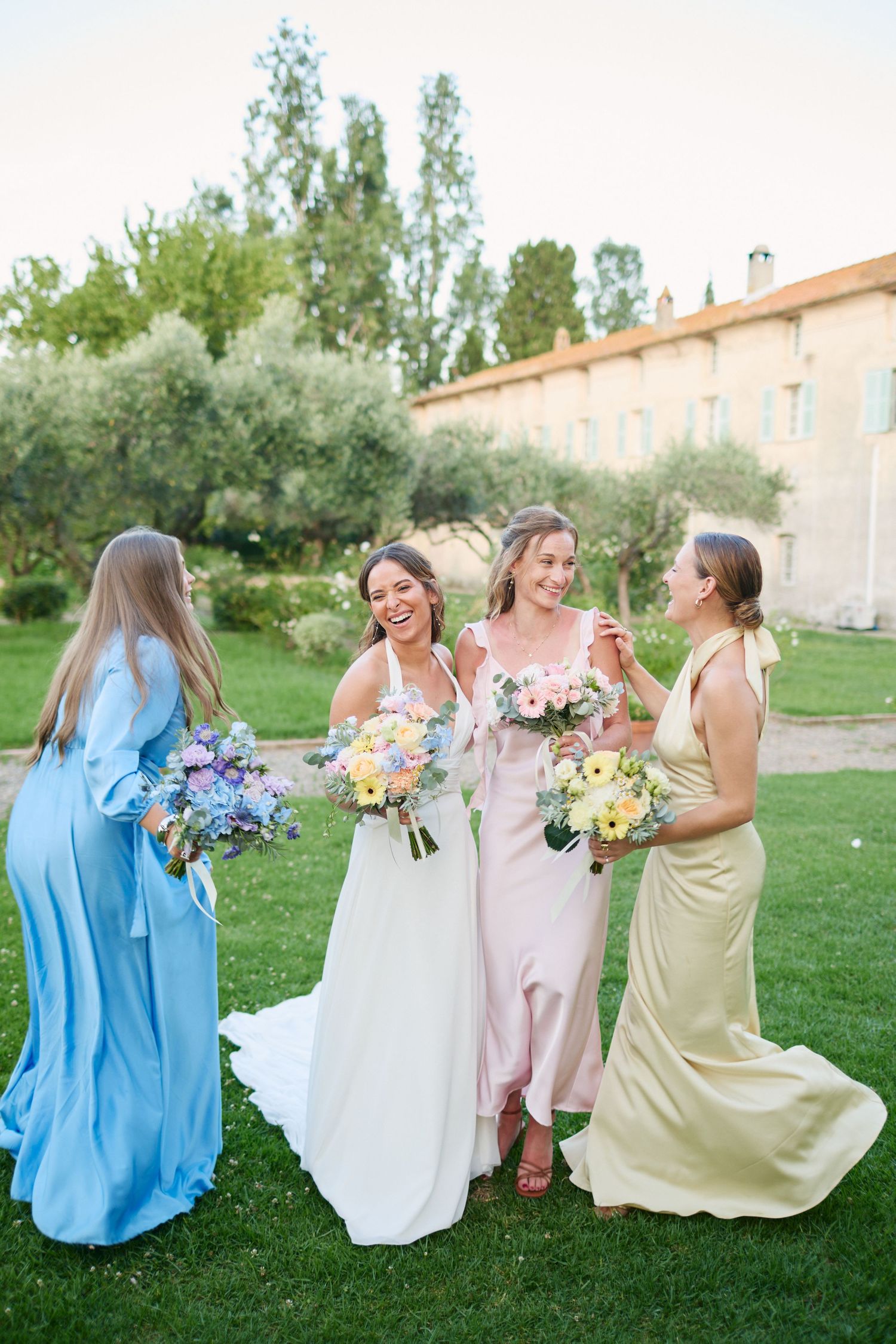 Bride laughing with bridesmaids in pastel gowns holding colorful bouquets on green lawn near stone building.