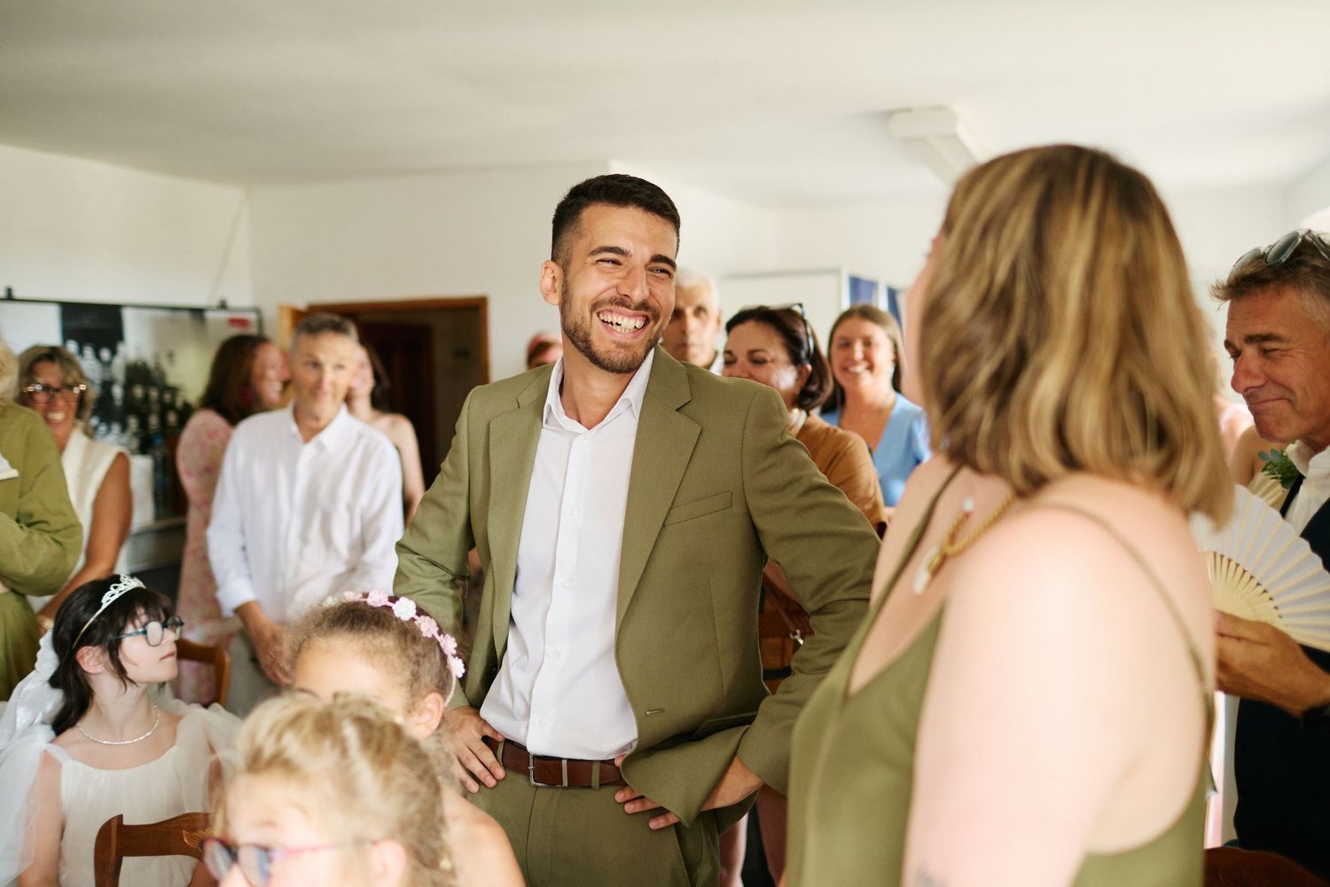 Smiling groomsman in olive suit mingles with guests at elegant wedding celebration.
