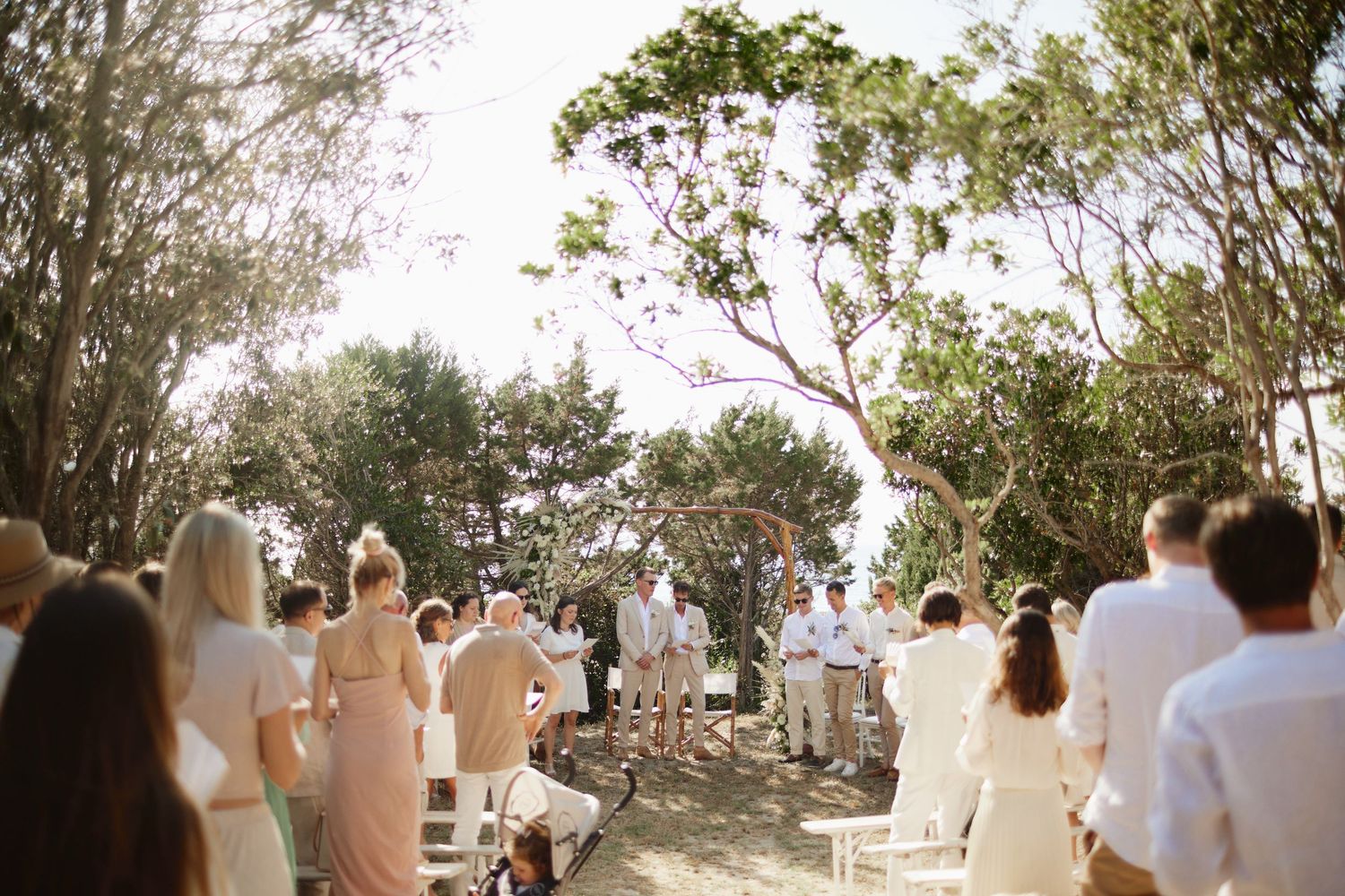 Guests stand and gather at a sunlit outdoor wedding ceremony lined with white chairs and trees.