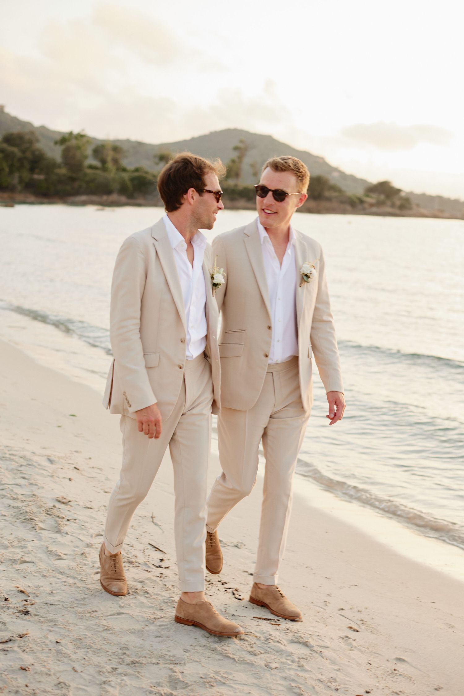 Two grooms in tan suits and sunglasses walking together along a tropical beach at sunset.