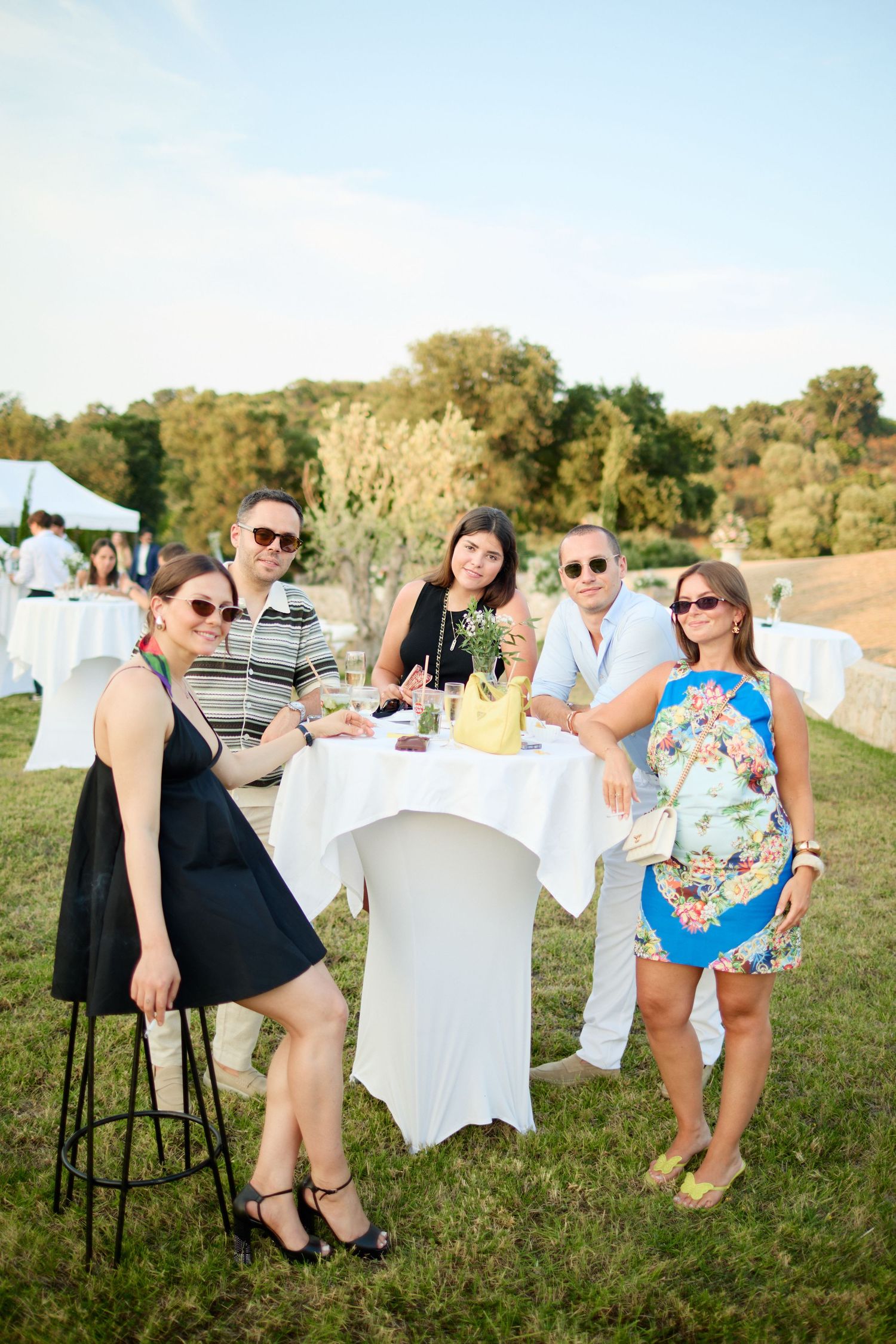 Five wedding guests smile at outdoor cocktail hour around white high-top table at sunset.