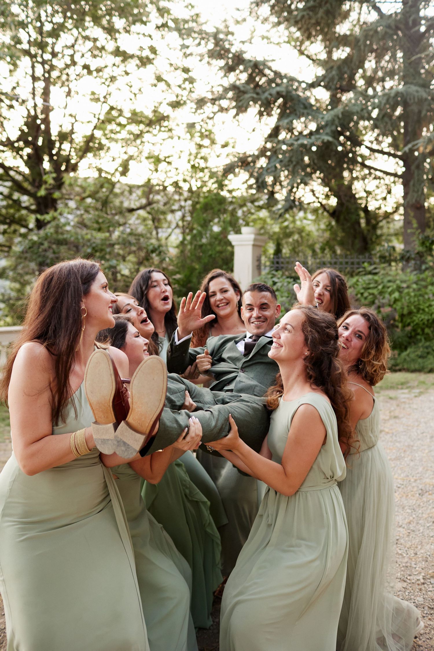 Bridesmaids in sage green dresses playfully lift grinning groom at outdoor wedding.