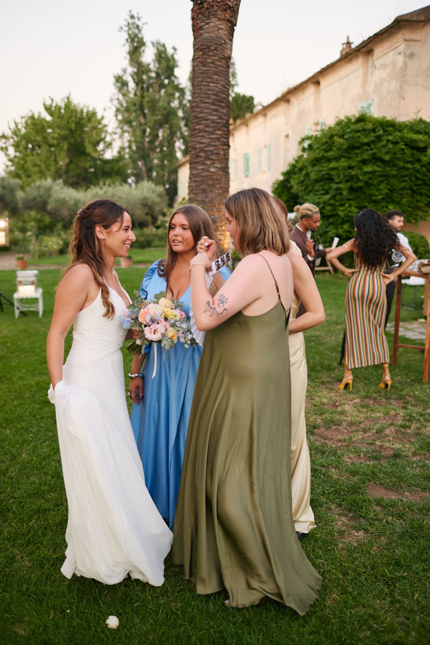 Bride in white gown chatting with guests in blue and green dresses at outdoor wedding.