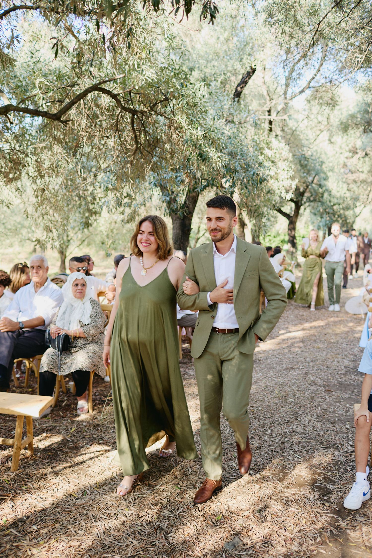 Smiling couple in coordinating olive green outfits walking down the outdoor wedding ceremony aisle together.