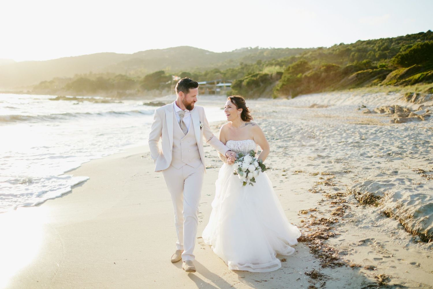 Couple de mariés marchant sur la plage au coucher du soleil après leur mariage à la Paillote Chez Francis en Corse.