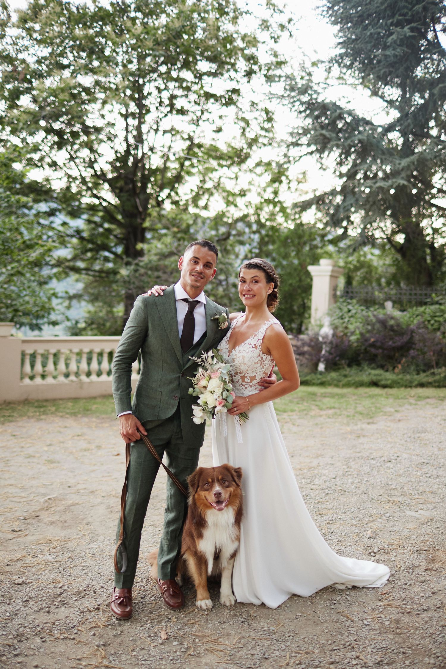 Bride and groom smile with their brown Australian Shepherd dog at outdoor wedding.