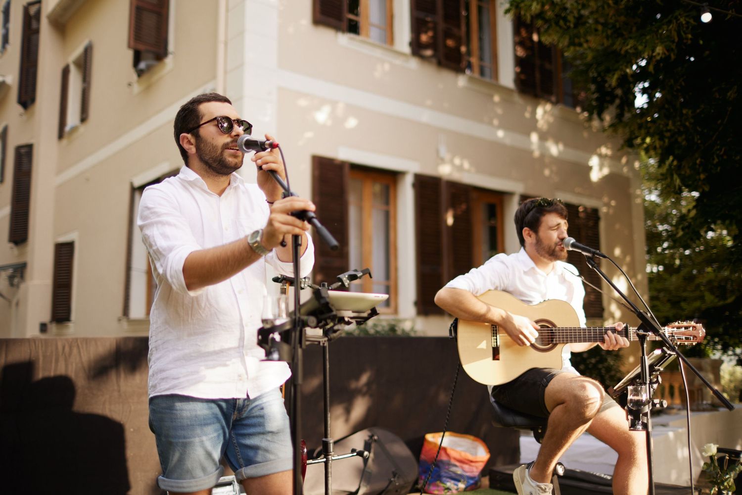 Two musicians perform outdoors, one singing into microphone, other playing acoustic guitar.