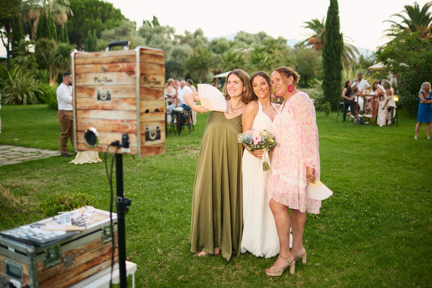 Bride posing with guests near a vintage photo booth at an outdoor garden wedding reception.