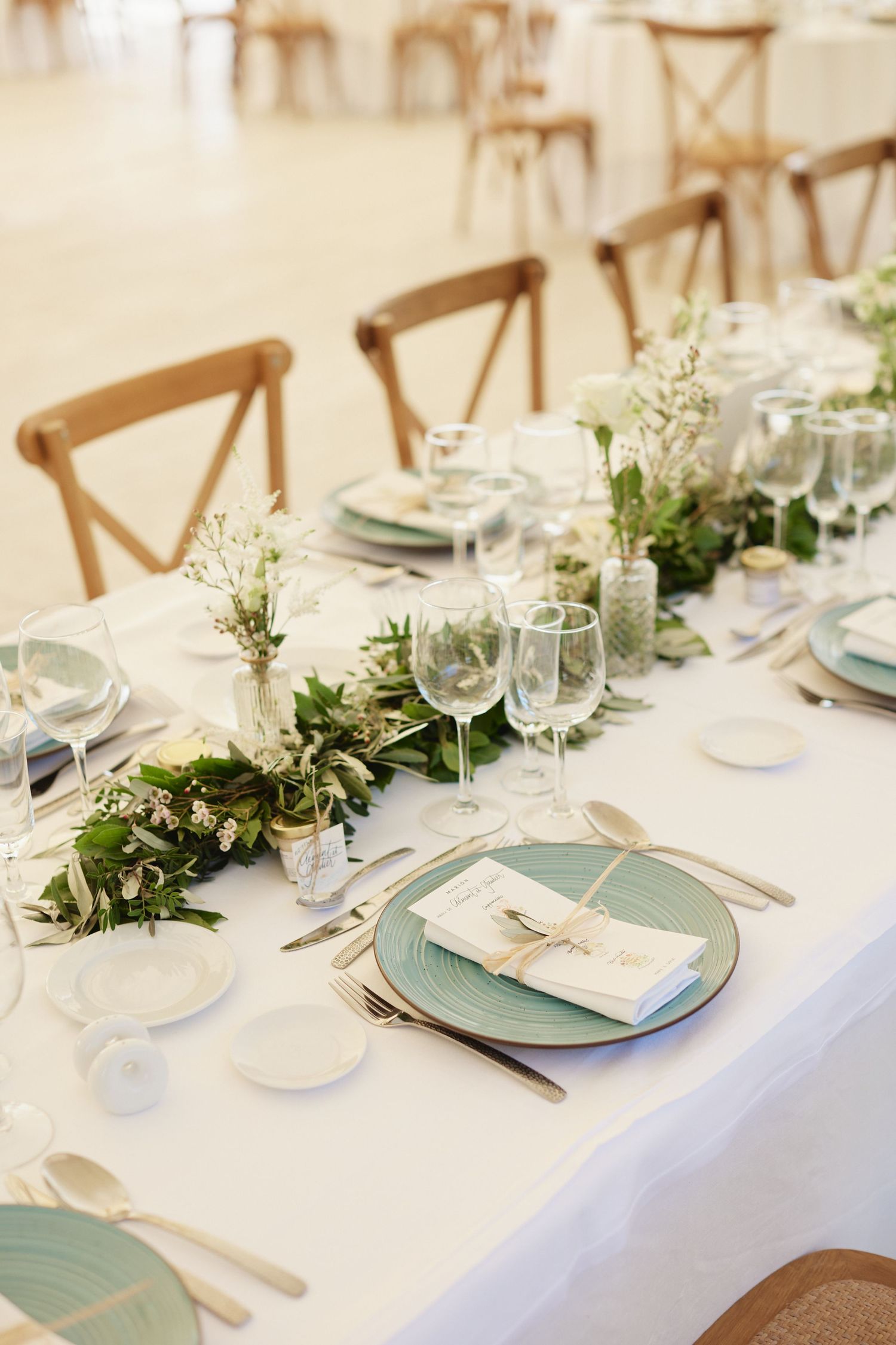 Elegant wedding reception table with green floral garland, sage charger plates, crystal glassware, and white linen.