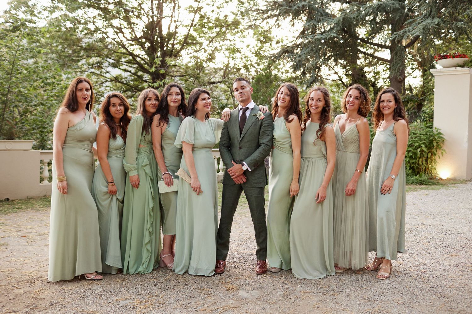 Groom in green suit stands with eight bridesmaids in sage green dresses outdoors.