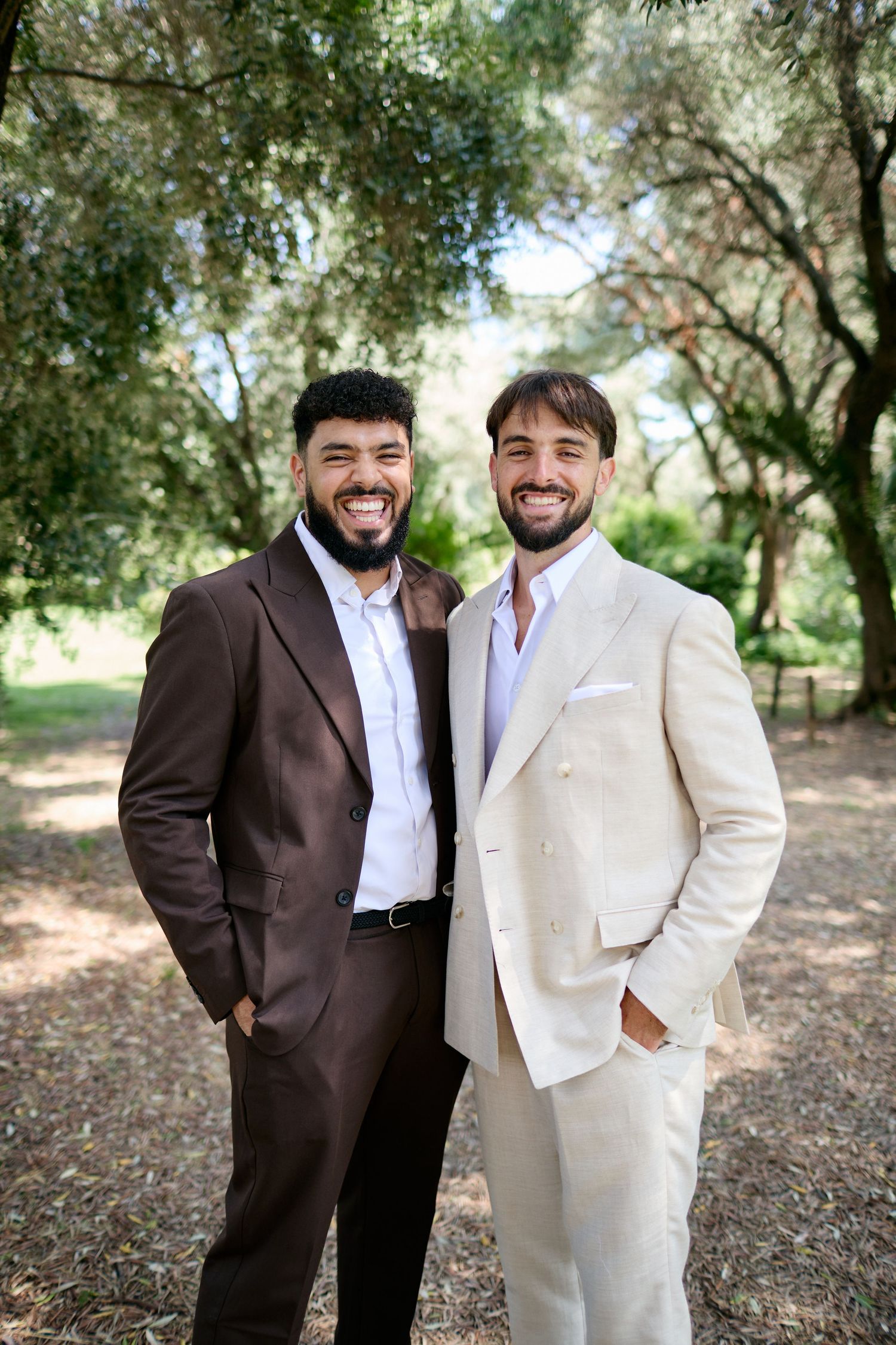 Two grooms smiling in a sunny park, one in brown suit and one in cream double-breasted suit.