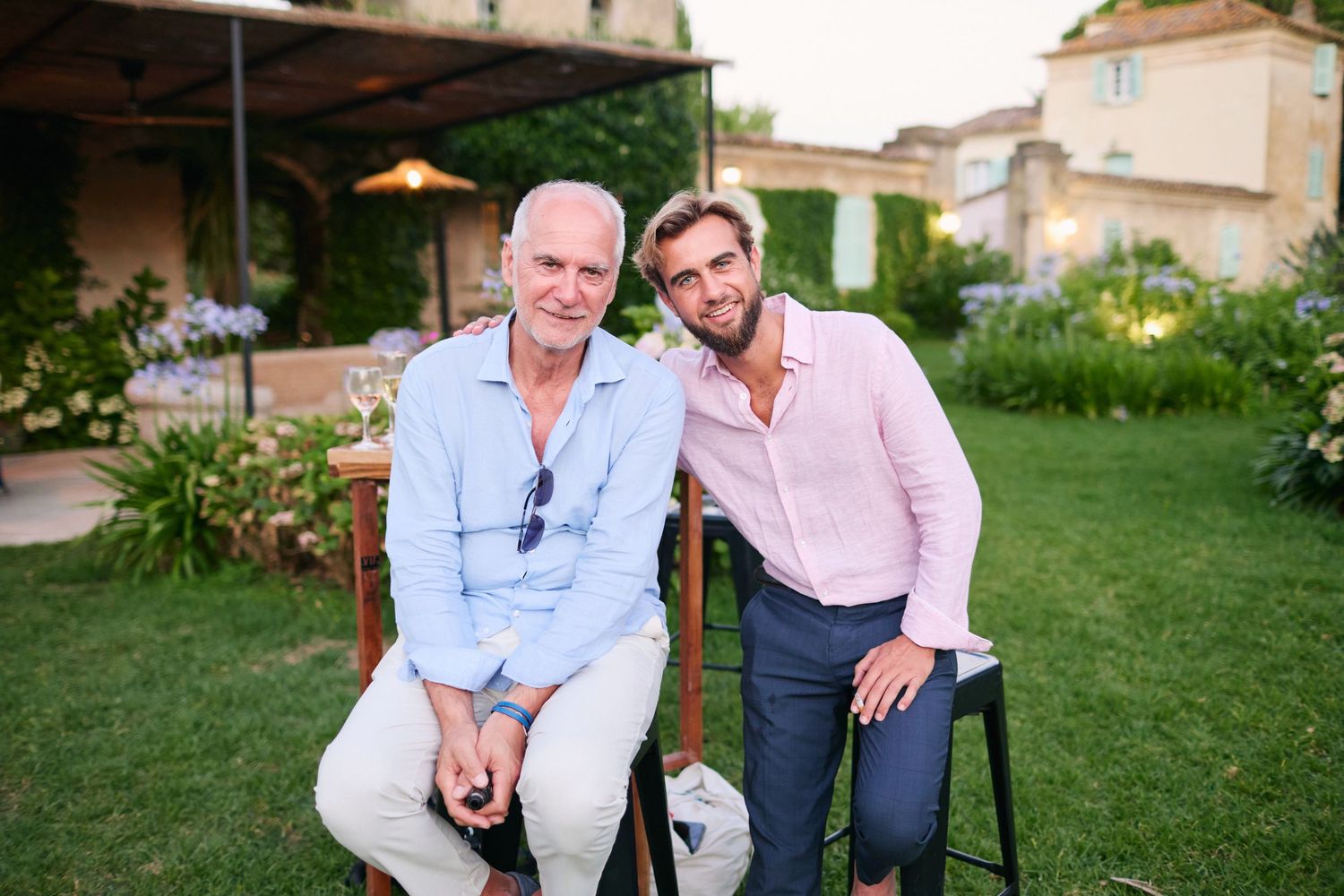 Two men in casual elegant attire sitting together at an outdoor wedding garden reception.