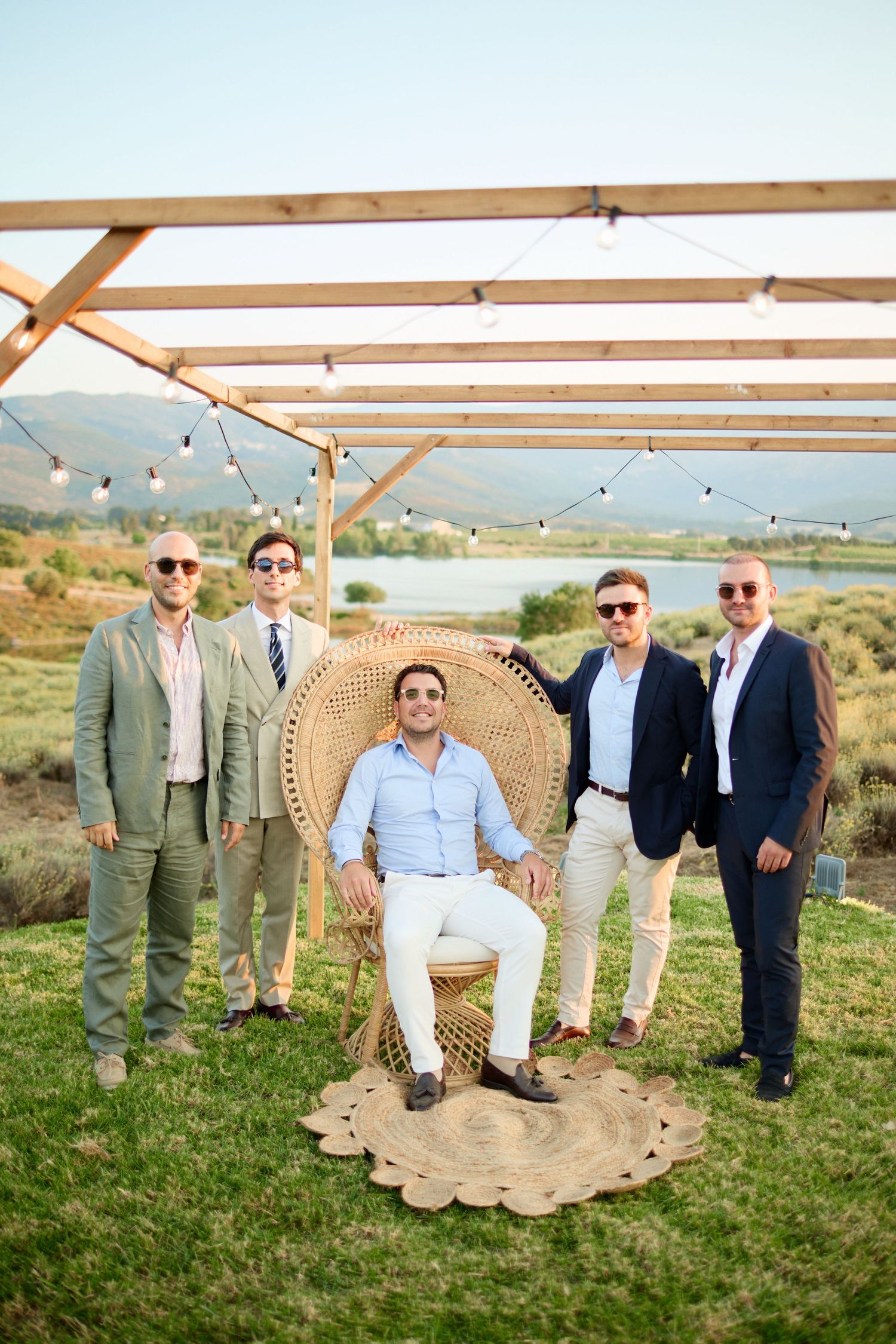 Five groomsmen posing under wooden pergola with string lights and lake view backdrop.