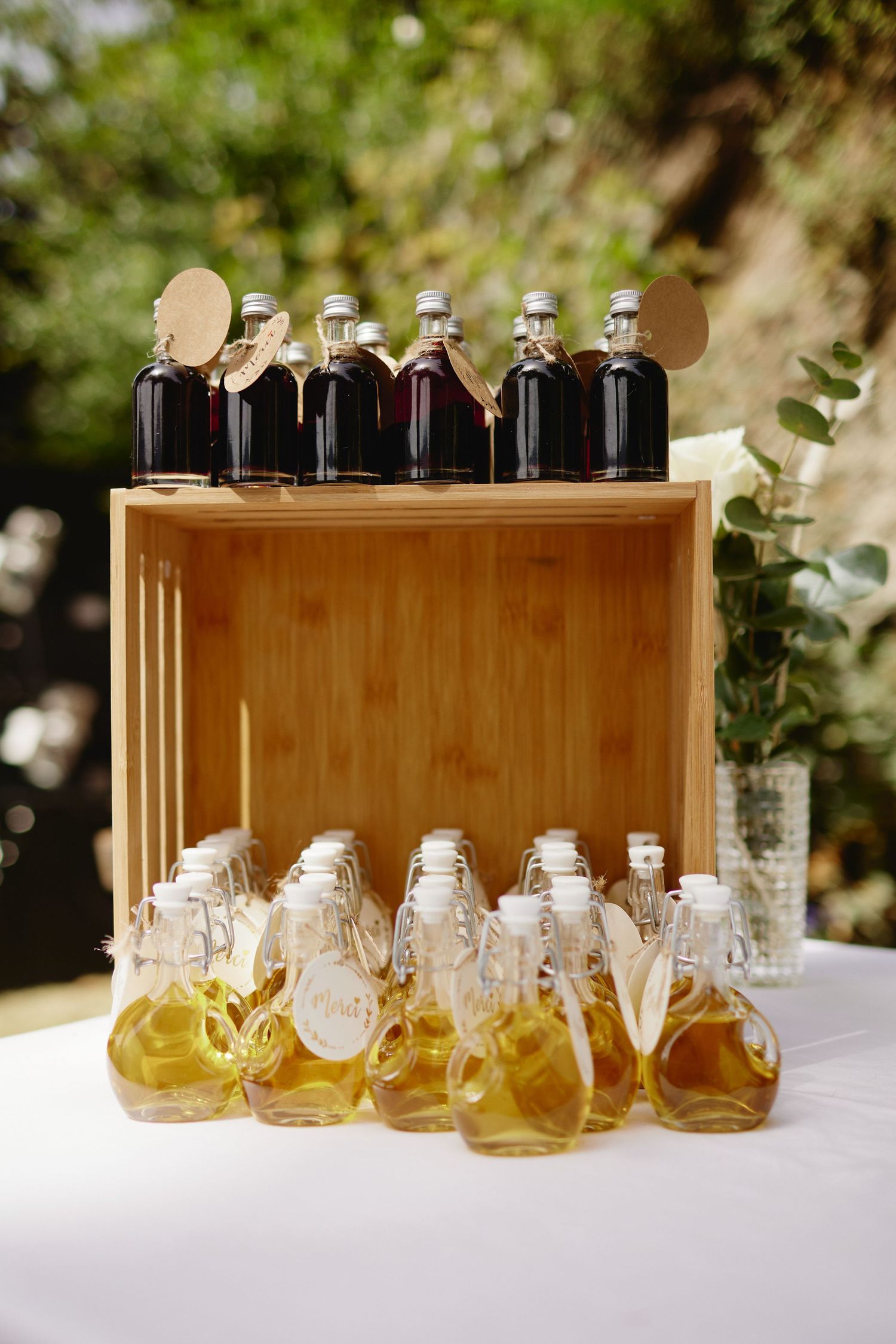 Wedding favor display featuring honey bear bottles and dark liquor bottles arranged on a wooden crate outdoors.