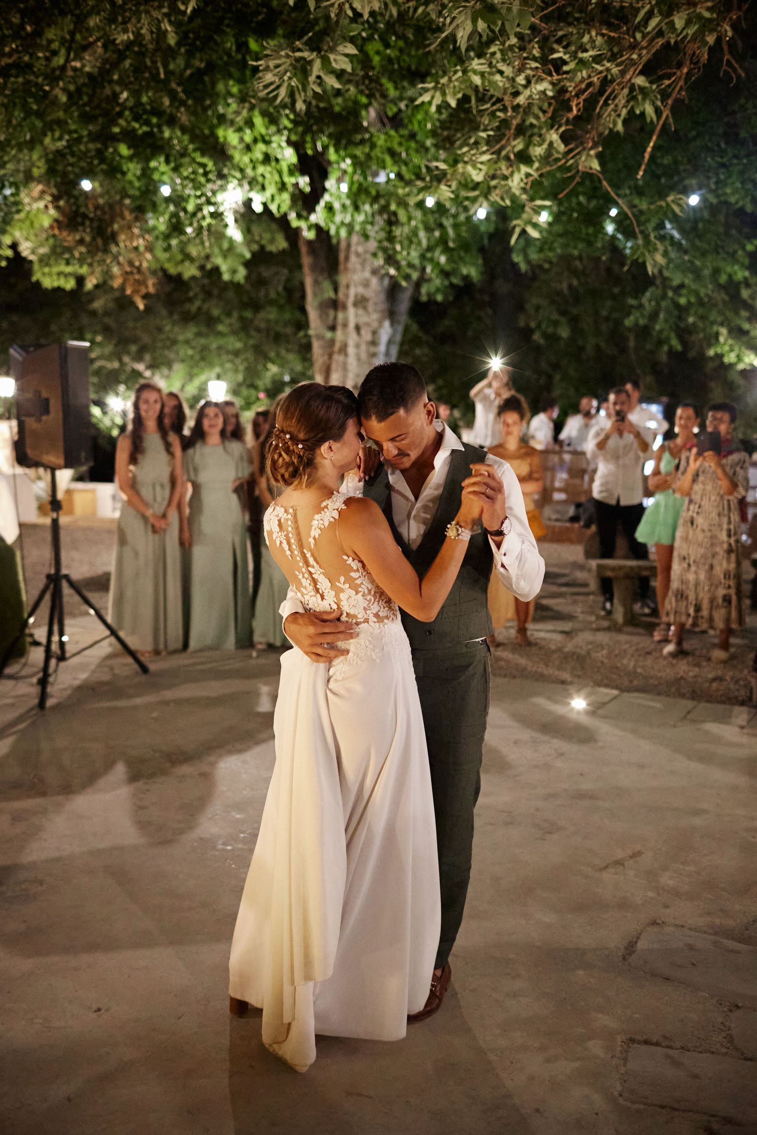 Bride and groom share their first dance under twinkling lights as wedding guests watch in outdoor evening reception.
