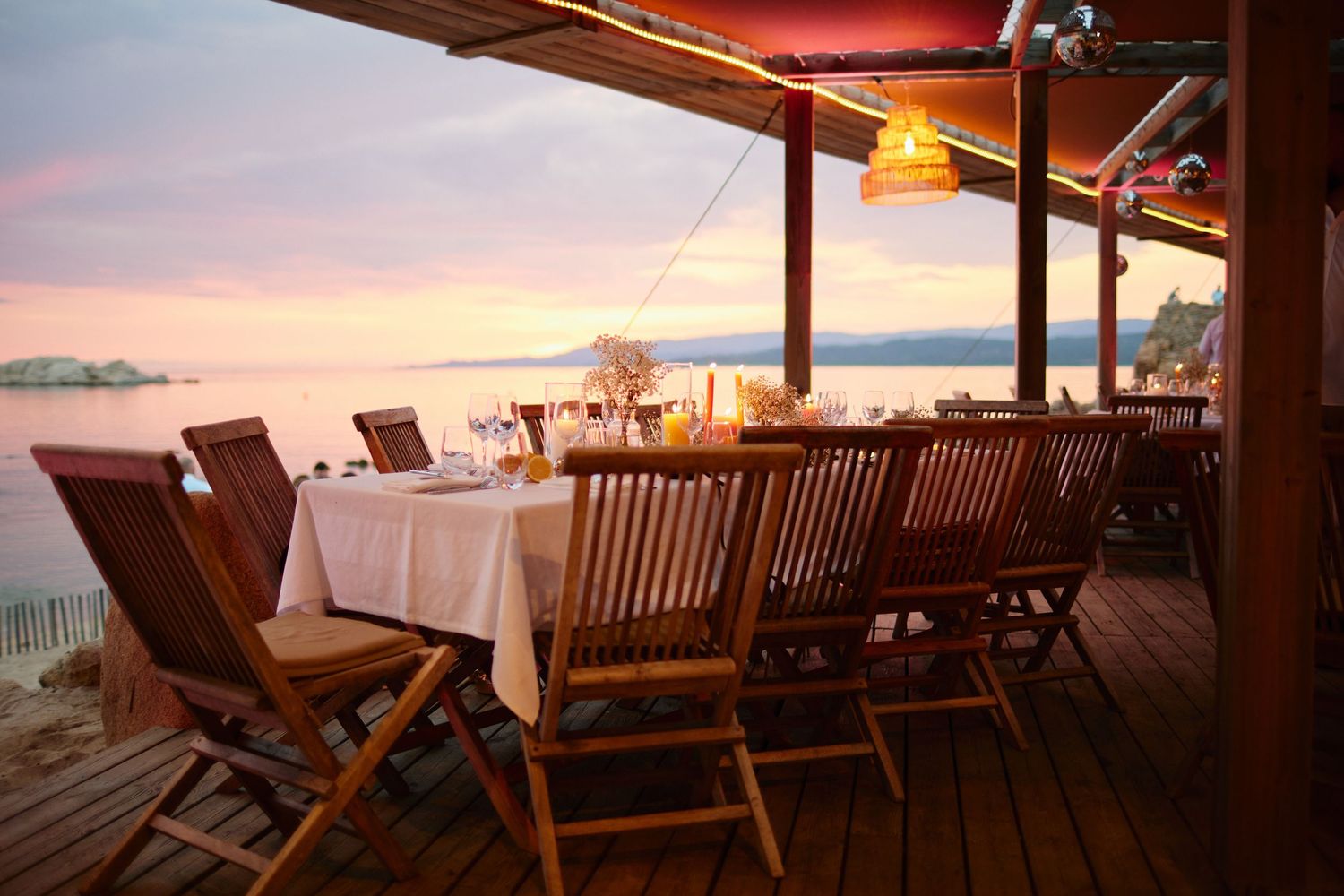 Elegant beachside restaurant terrace with wooden chairs and tables set for a romantic dinner at sunset.