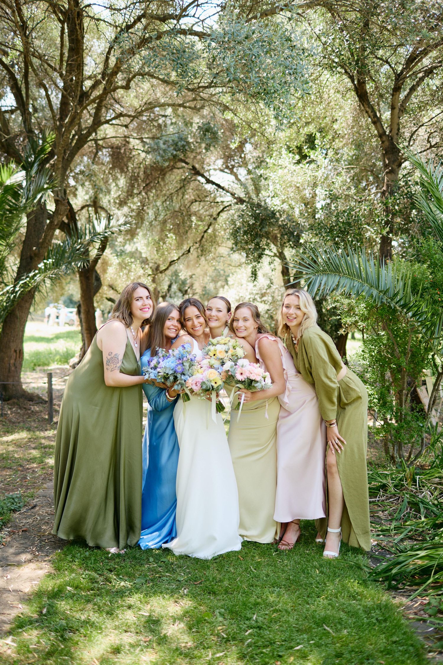 Bride in white gown hugs five bridesmaids in colorful mismatched dresses holding bouquets in garden setting.
