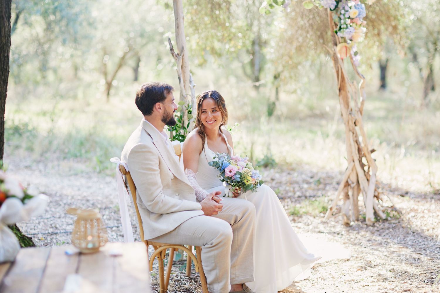 Happy bride and groom seated together in a sunlit olive grove during their outdoor wedding ceremony.