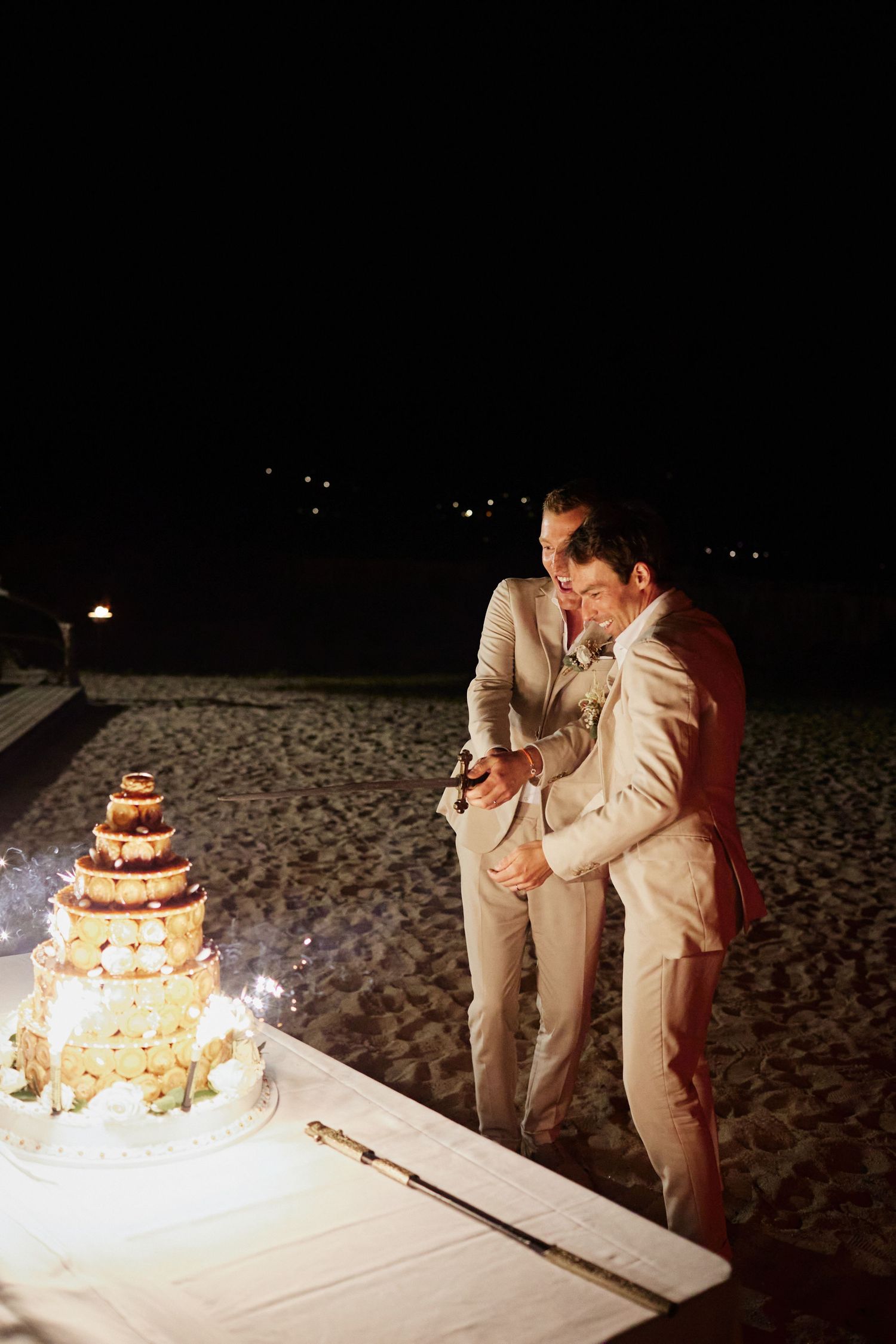 Two grooms in beige suits cutting their multi-tiered wedding cake on the beach at night with sparklers.