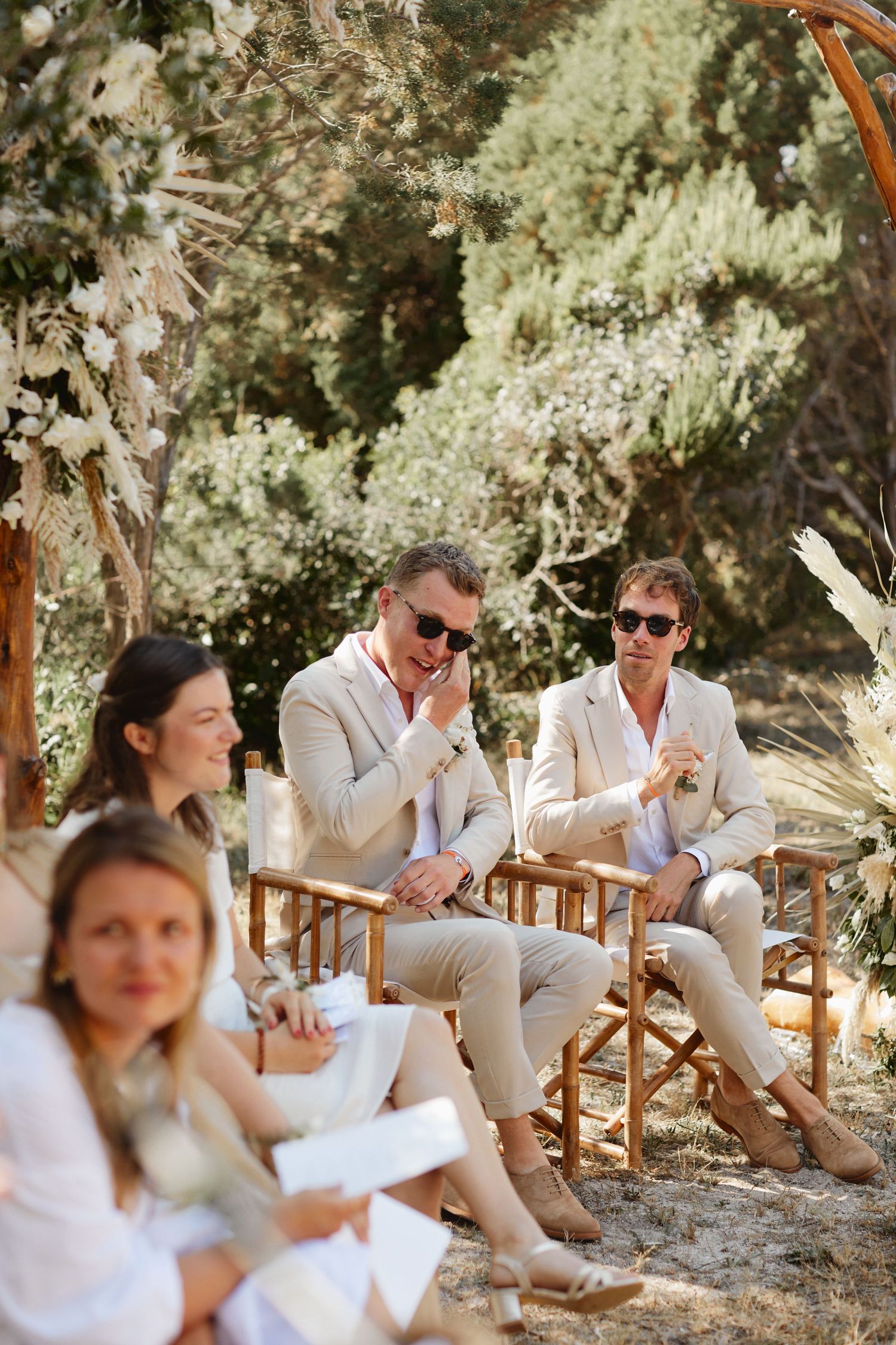 Two men in beige suits and sunglasses sit on wooden chairs at an outdoor wedding ceremony.