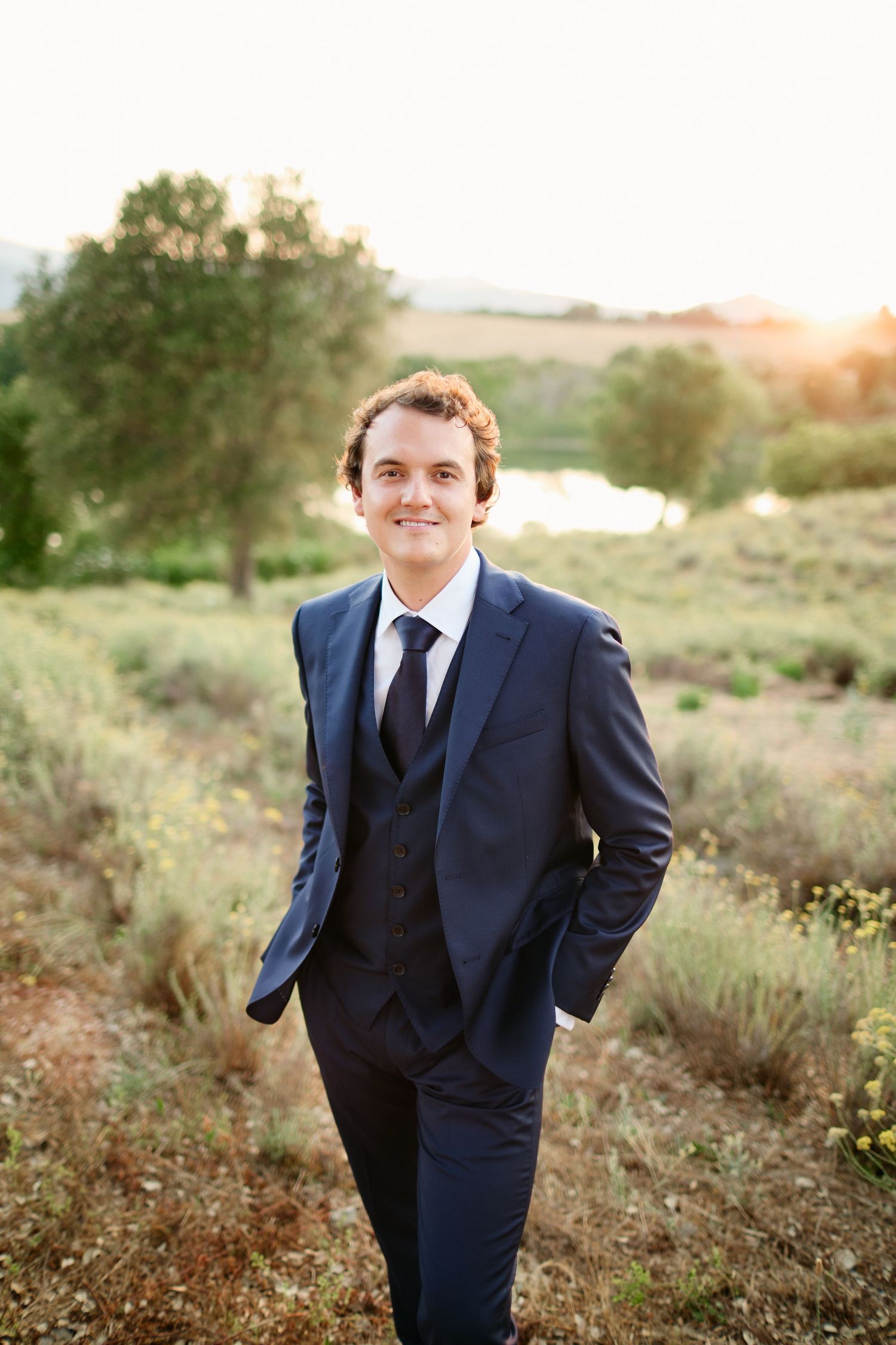 Handsome groom in navy blue suit smiles confidently in outdoor lavender field.