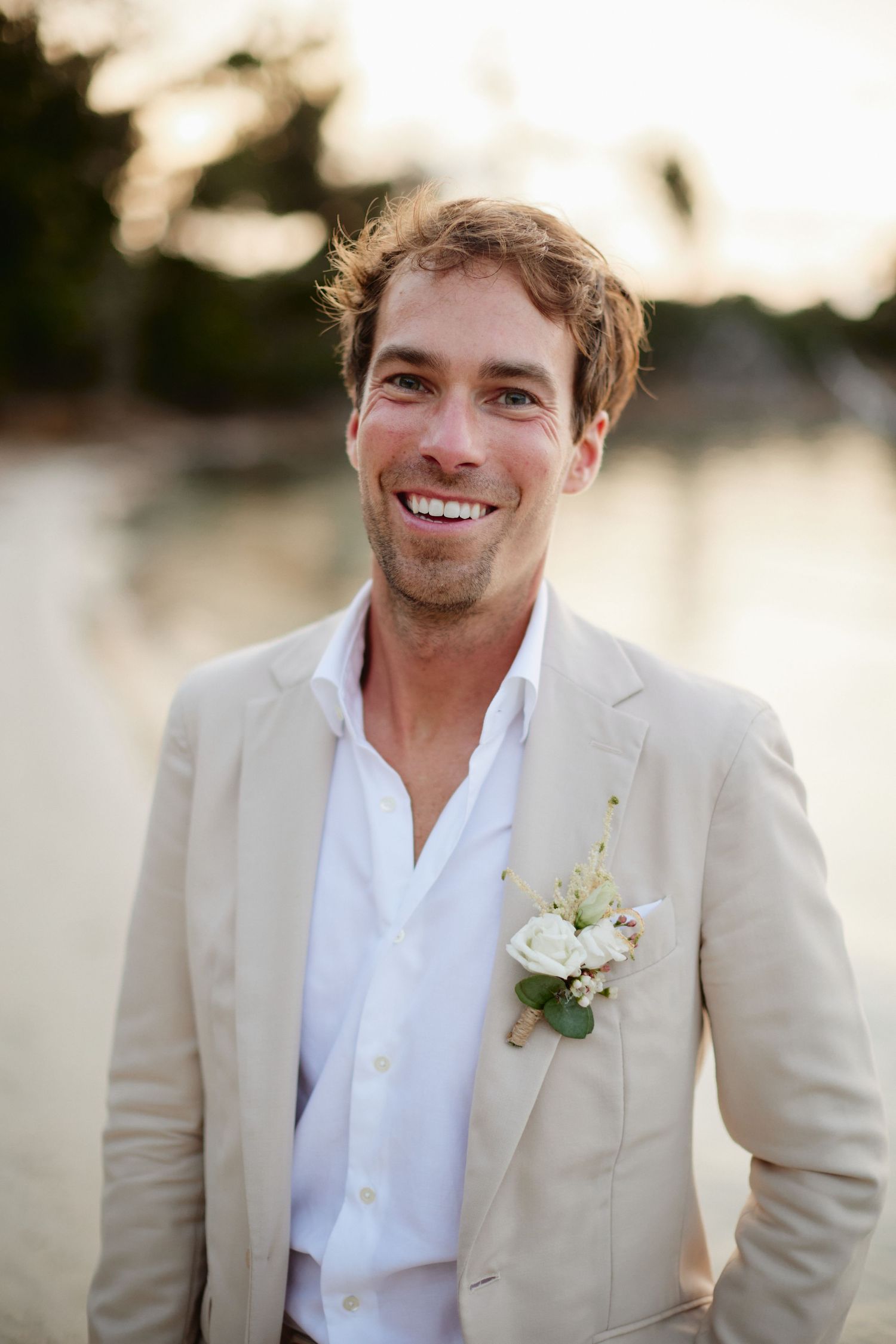 Smiling groom in beige blazer with white floral boutonniere at a tropical beach wedding.