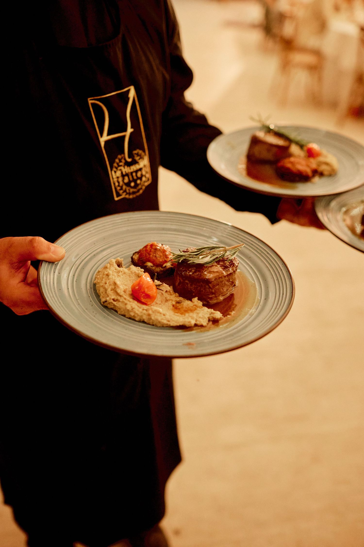 Waiter in black uniform serving two plated filet mignon dishes with mashed potatoes at a wedding reception.