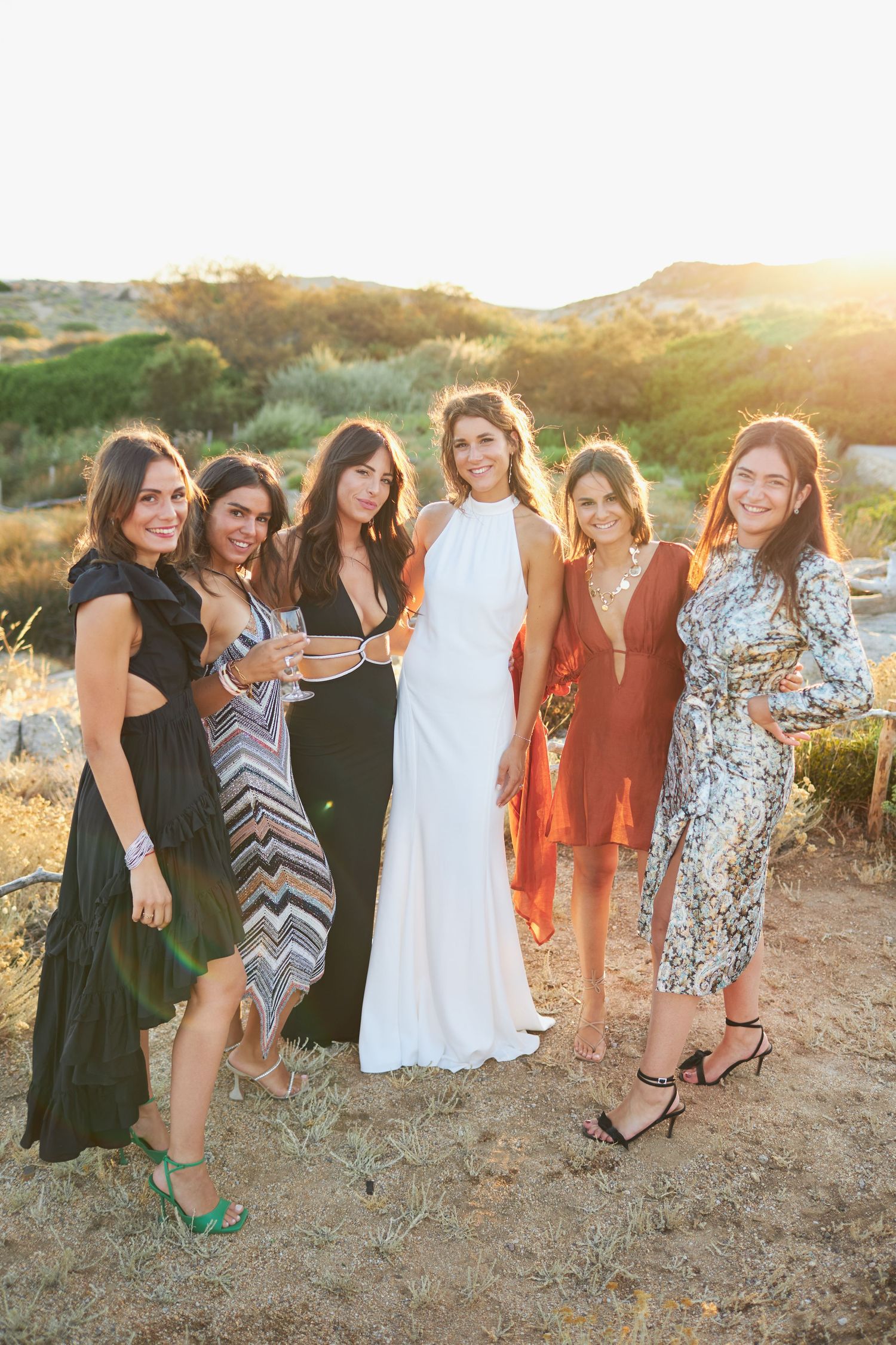 Bride in white halter gown posing with six female guests at a sun-drenched outdoor wedding.