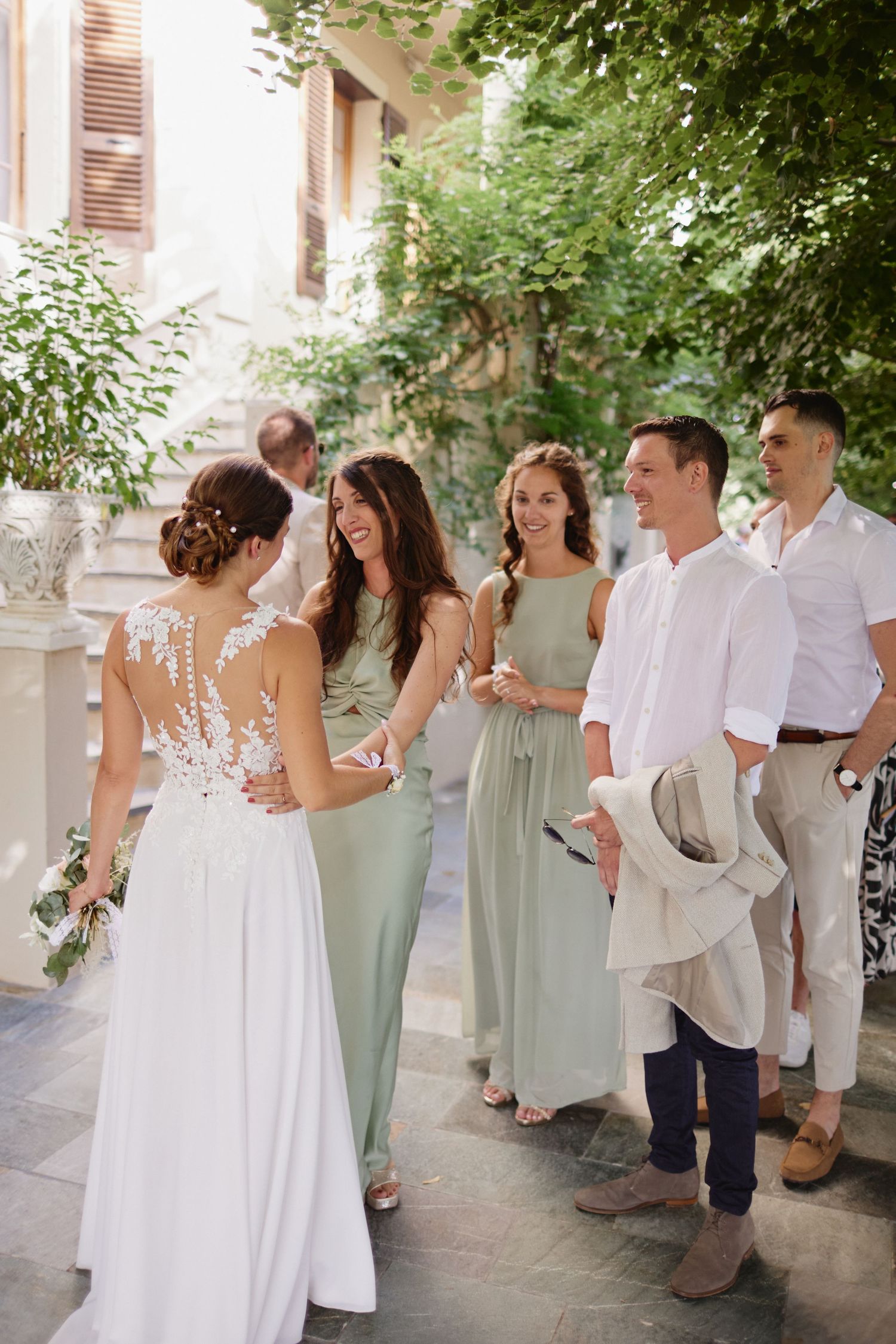 Bride in lace wedding dress greeting bridesmaids in sage green gowns at outdoor venue.