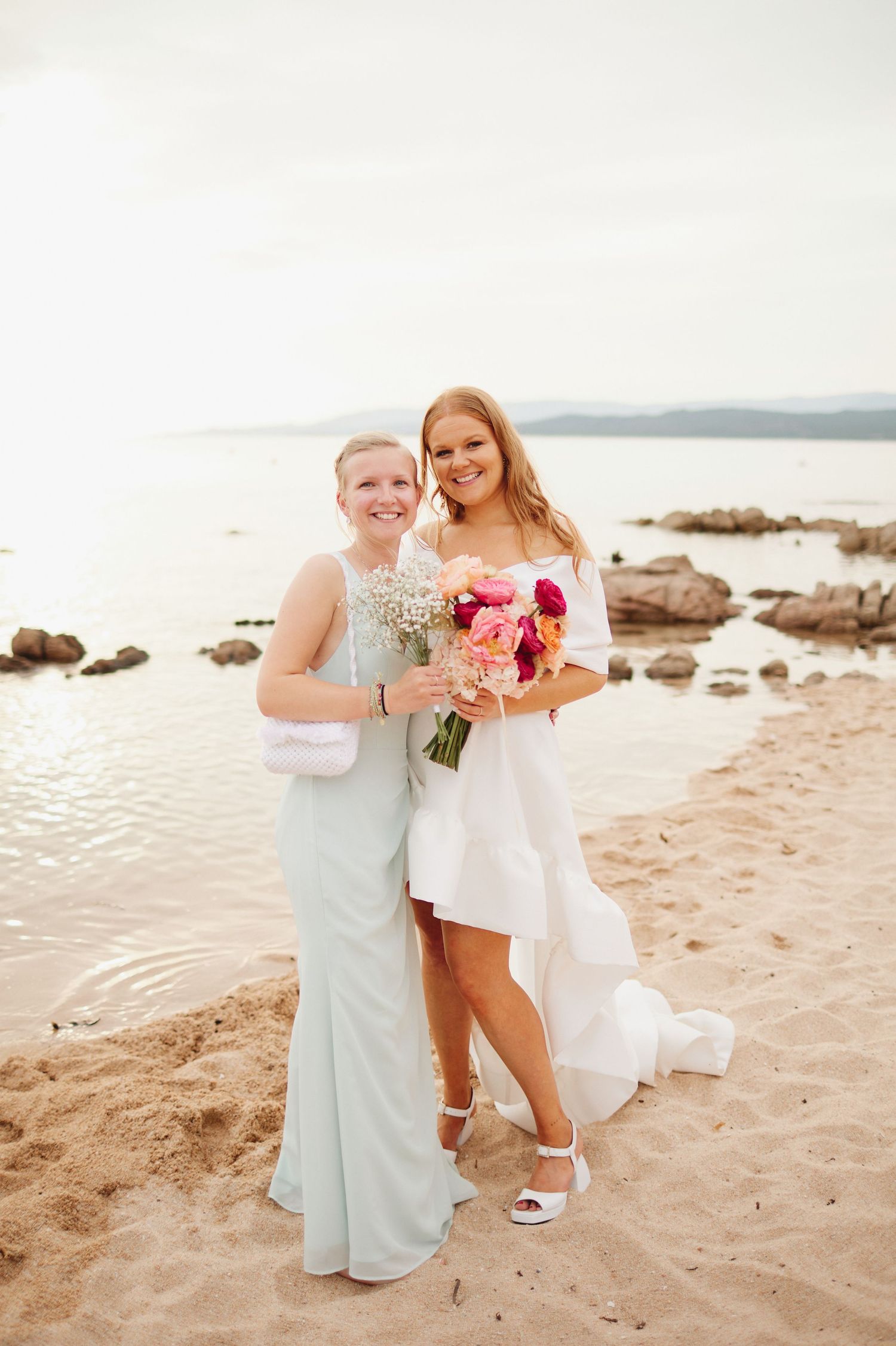 Bride in white mini dress holding pink bouquet posing with bridesmaid in mint gown on beach.