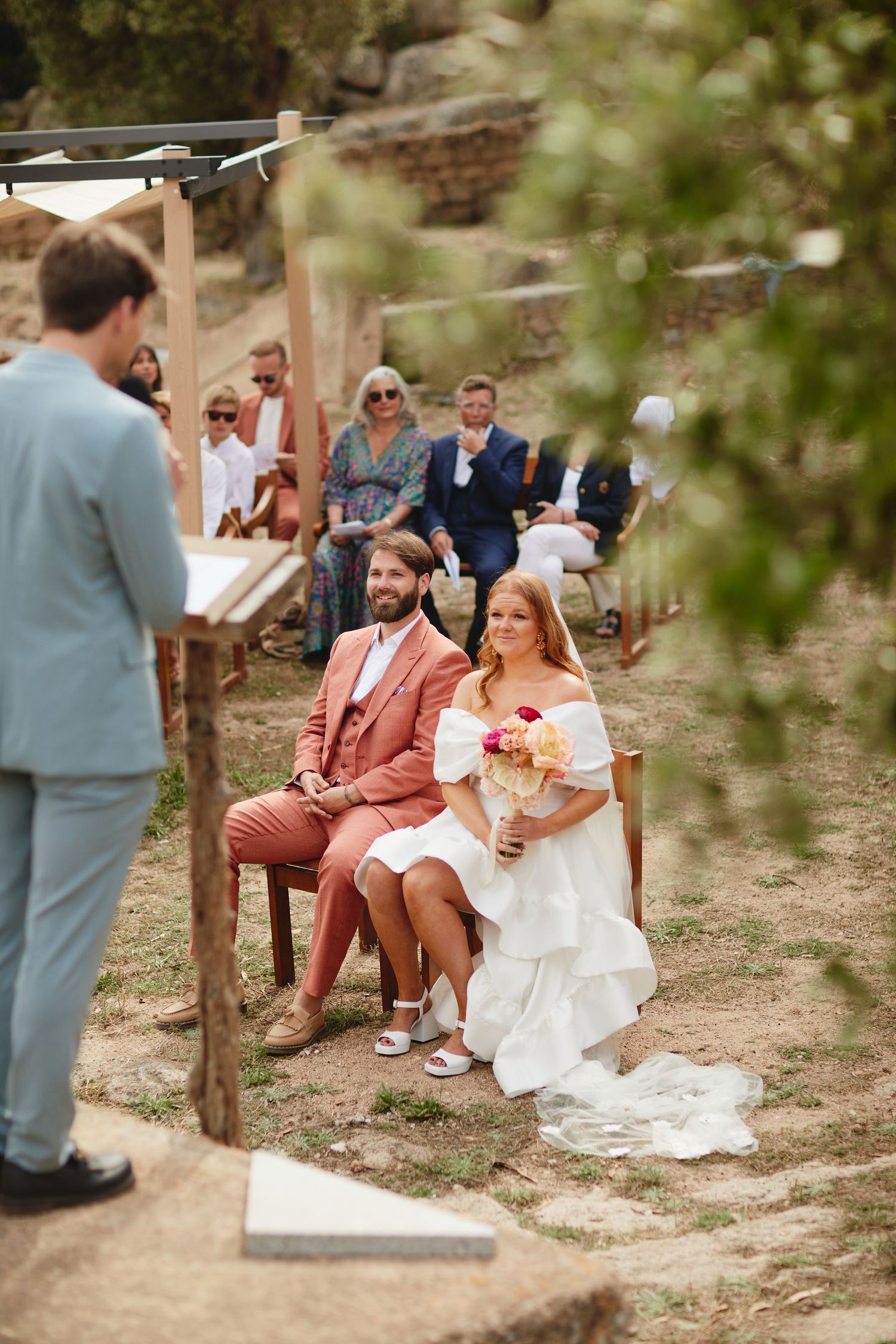 Bride and groom in coral suit sit together during outdoor wedding ceremony with guests behind.
