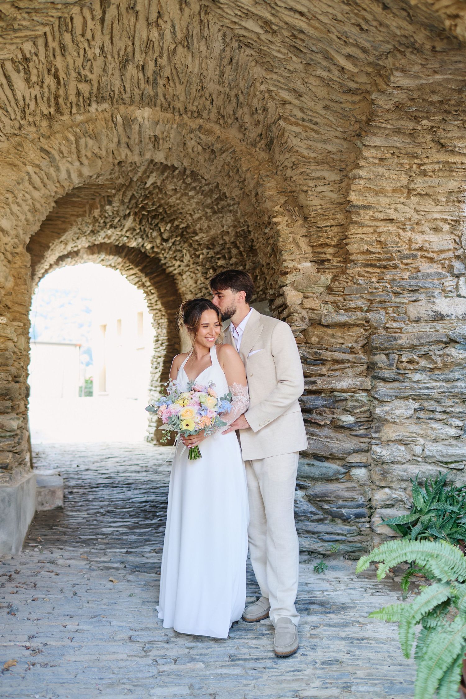Couple embraces tenderly under rustic stone arches holding pastel wedding bouquet.