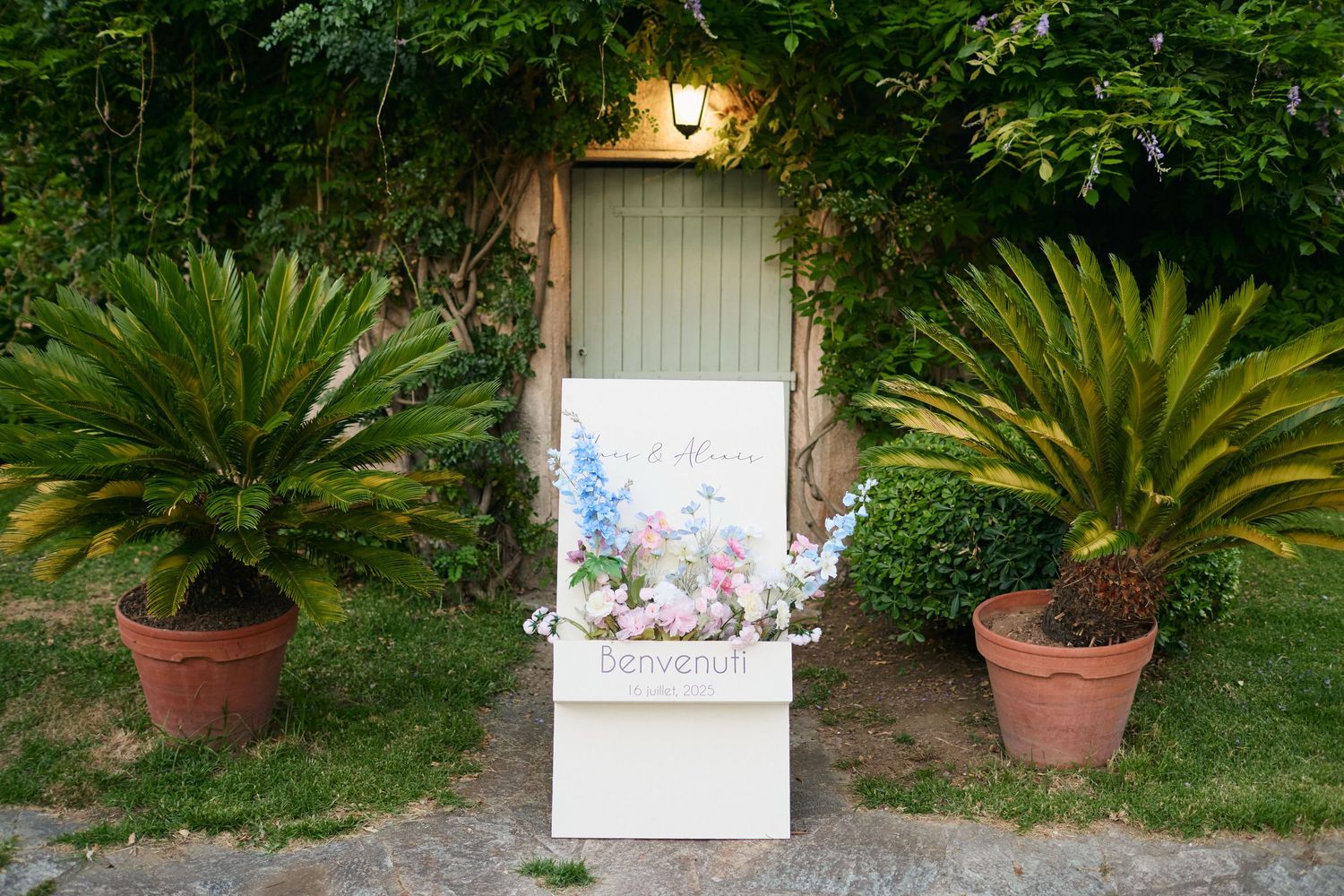 Elegant white welcome sign adorned with floral arrangement flanked by potted palms at wedding entrance.