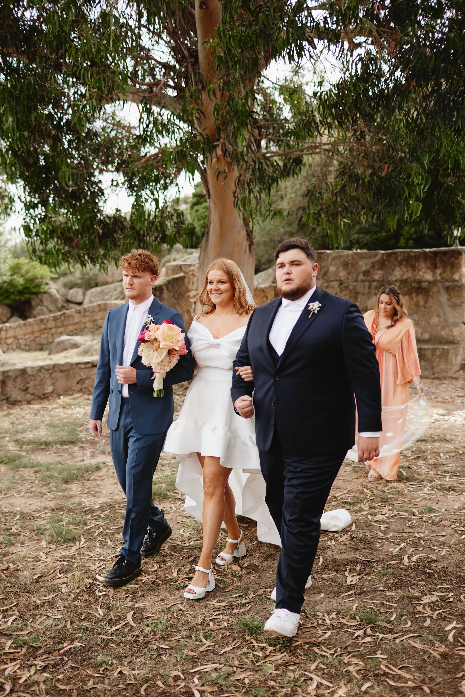 Bride in white dress walks with two groomsmen holding bouquet at outdoor rustic wedding.