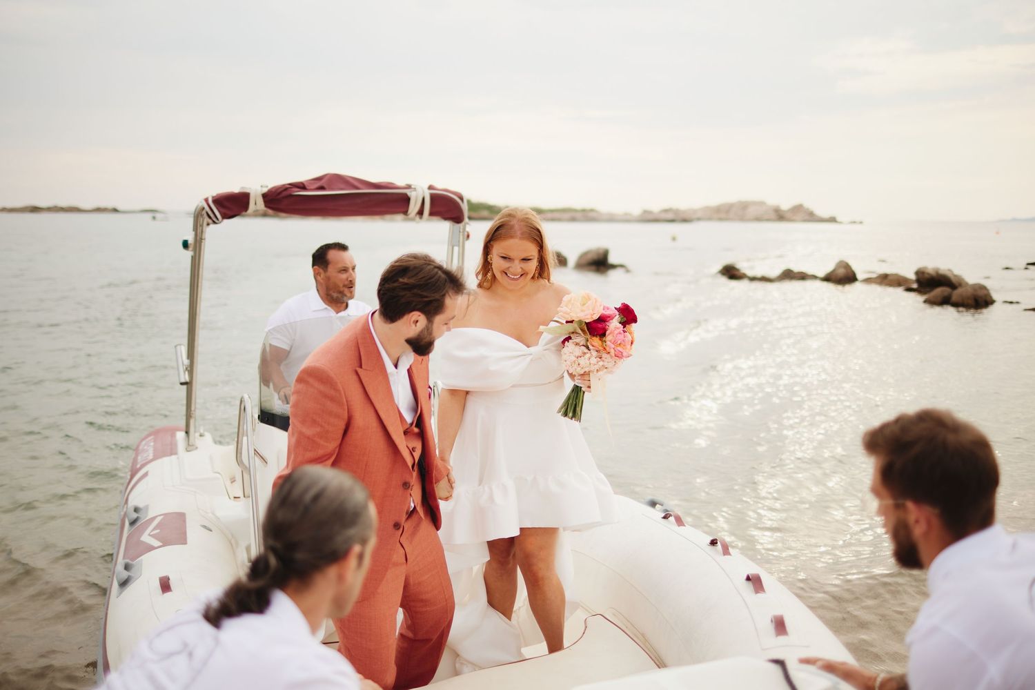 Smiling bride in white mini dress steps off a boat assisted by groom in coral suit.