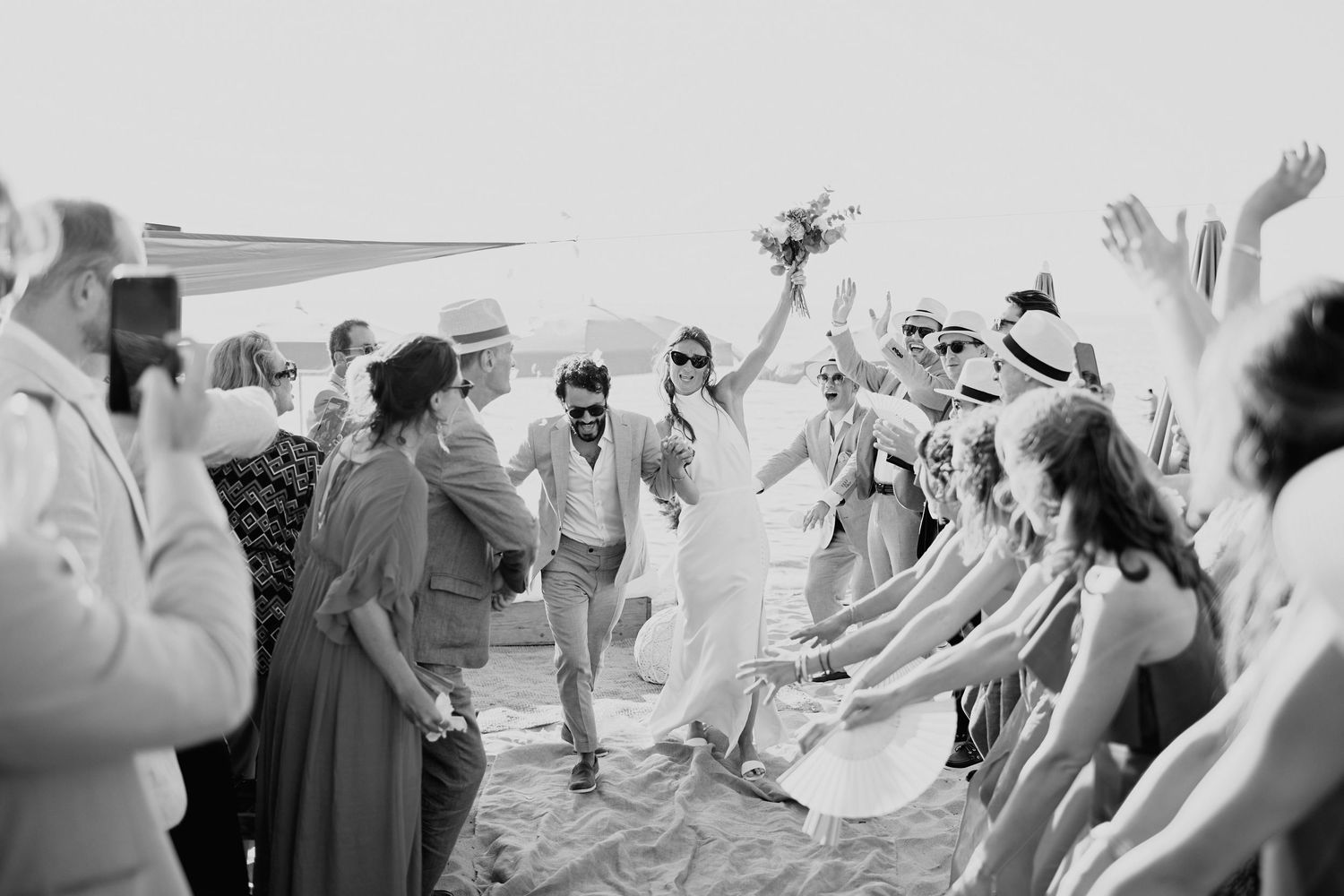 Black and white photo of newlyweds walking through cheering wedding guests on a sunny beach celebration.