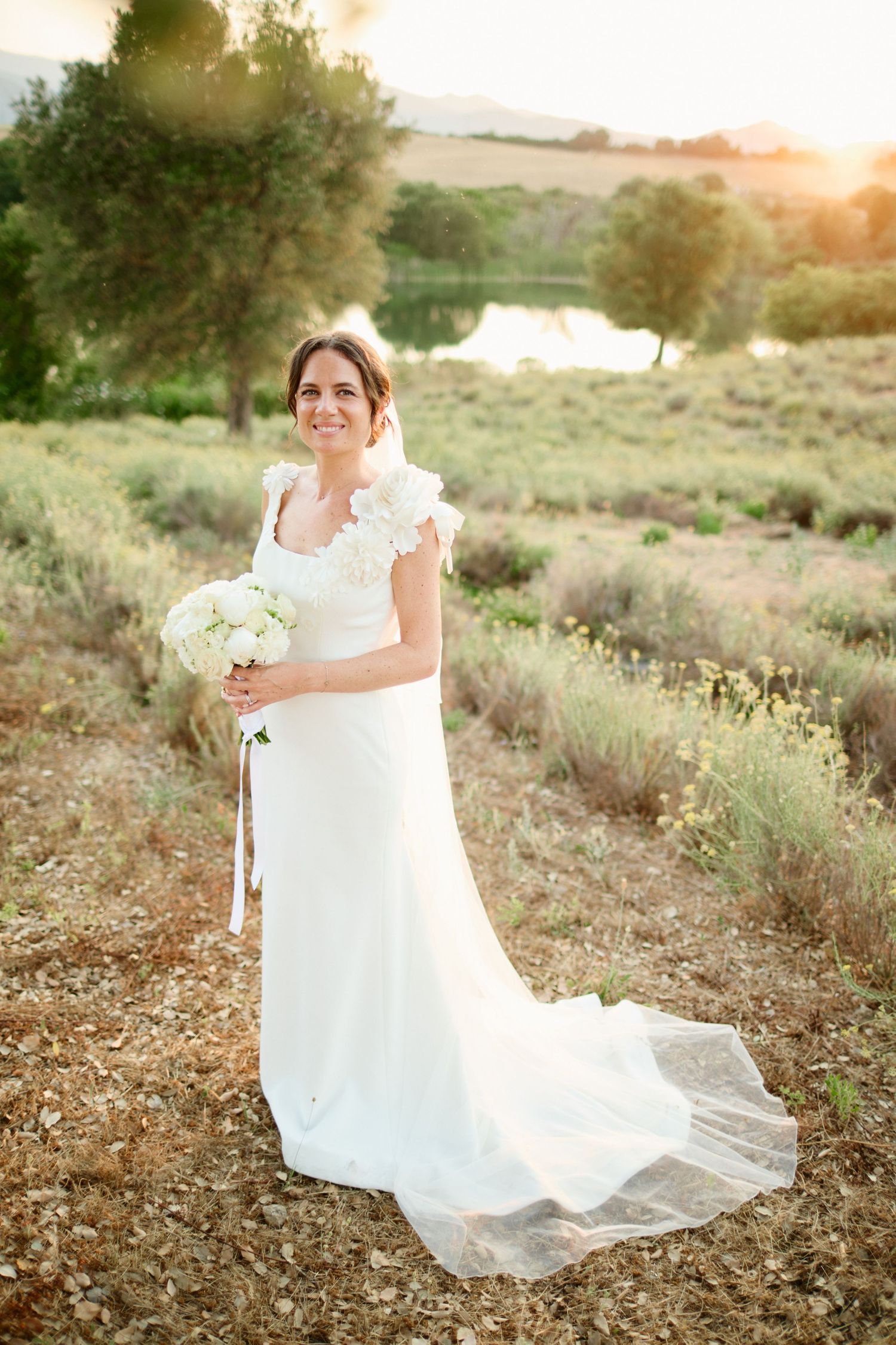 Elegant bride in white gown holds bouquet in sunlit lavender field at sunset.
