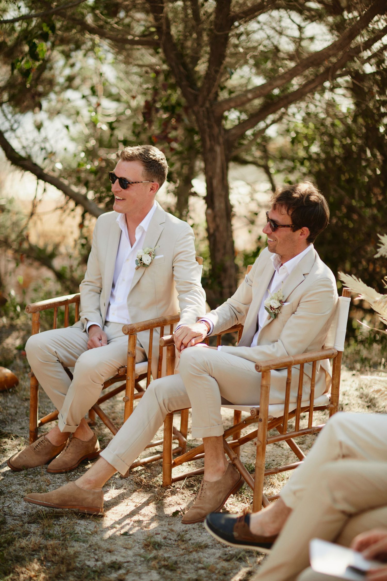 Two grooms in matching beige suits and sunglasses sit together at an outdoor wedding ceremony.