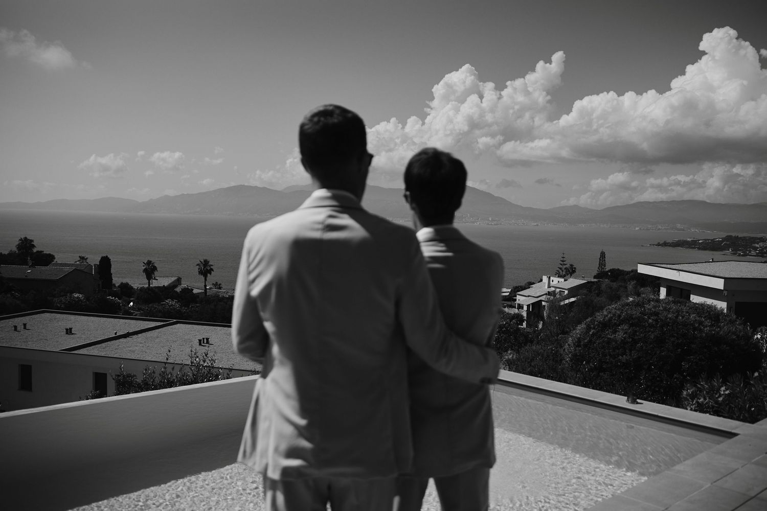 Two grooms in beige suits embracing while overlooking scenic coastal landscape.