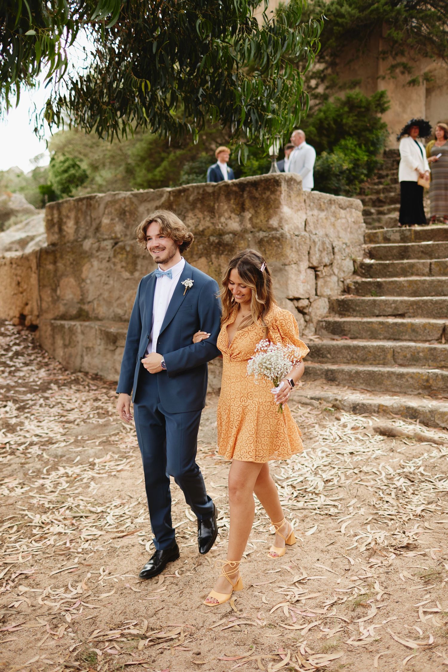 Smiling groomsman in blue suit escorts bridesmaid in yellow dress at outdoor wedding ceremony.