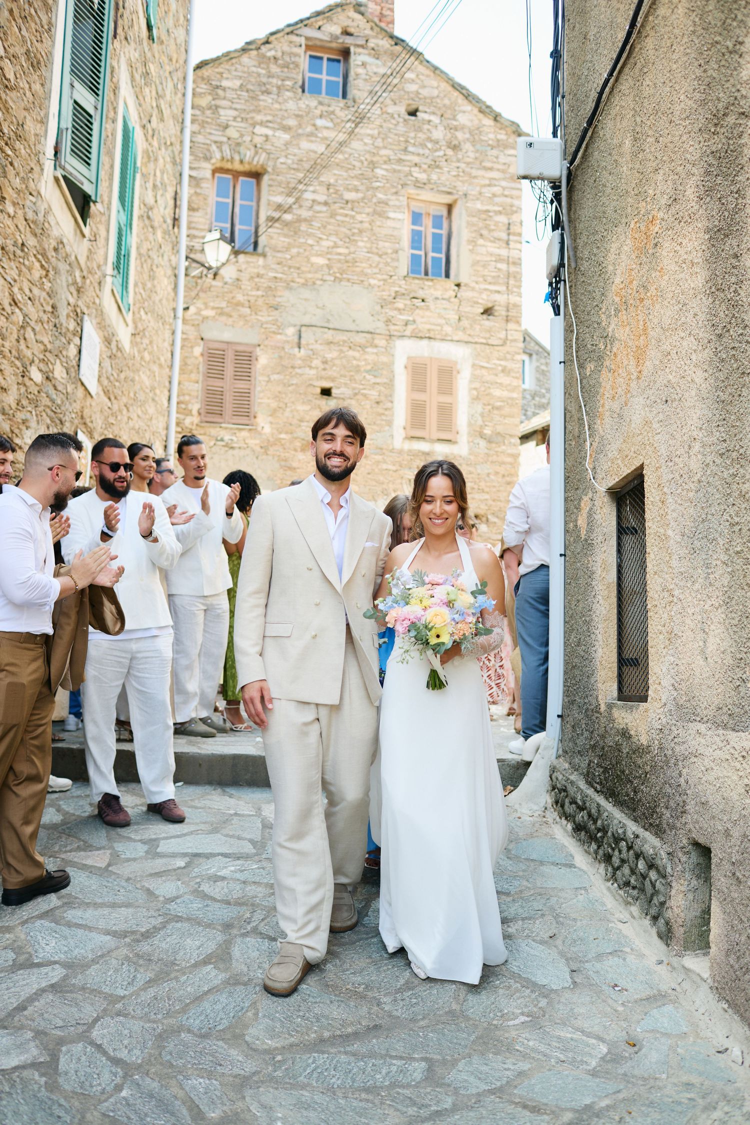 Smiling bride and groom exit ceremony through stone village street with cheering guests.